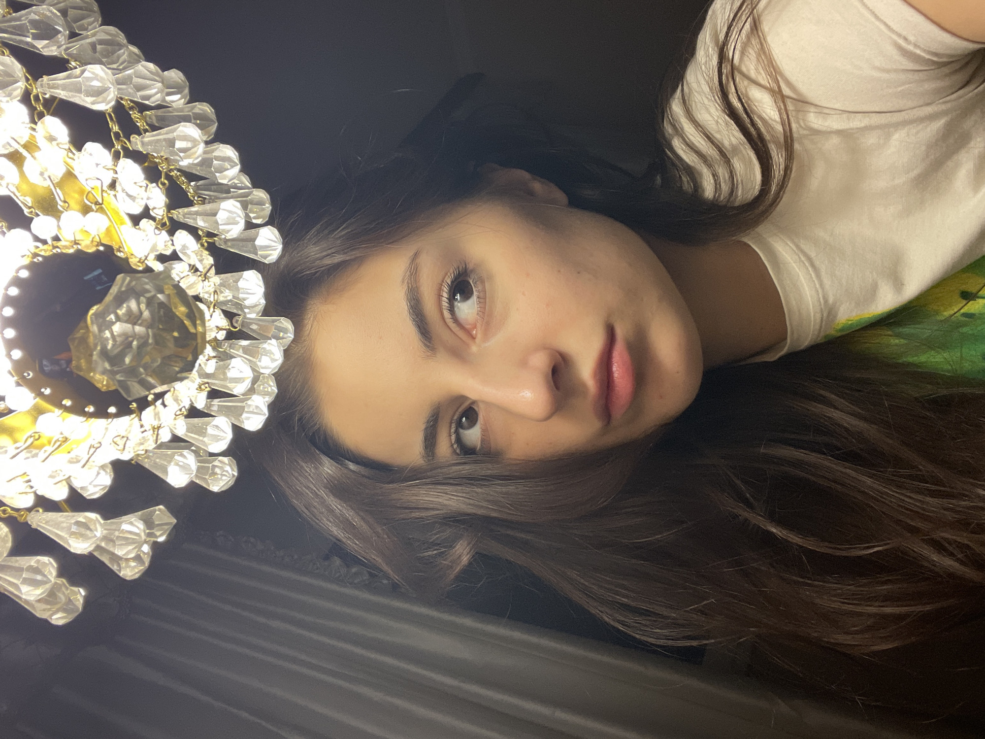 A young woman with long brown hair is lying down, face toward the camera. She is wearing a white t-shirt. Next to her head is a lit chandelier with multiple crystal pendants. The background appears to be a dark curtain or wall. There is no information in the image that allows for identification of the location as Moscow, Russia.
