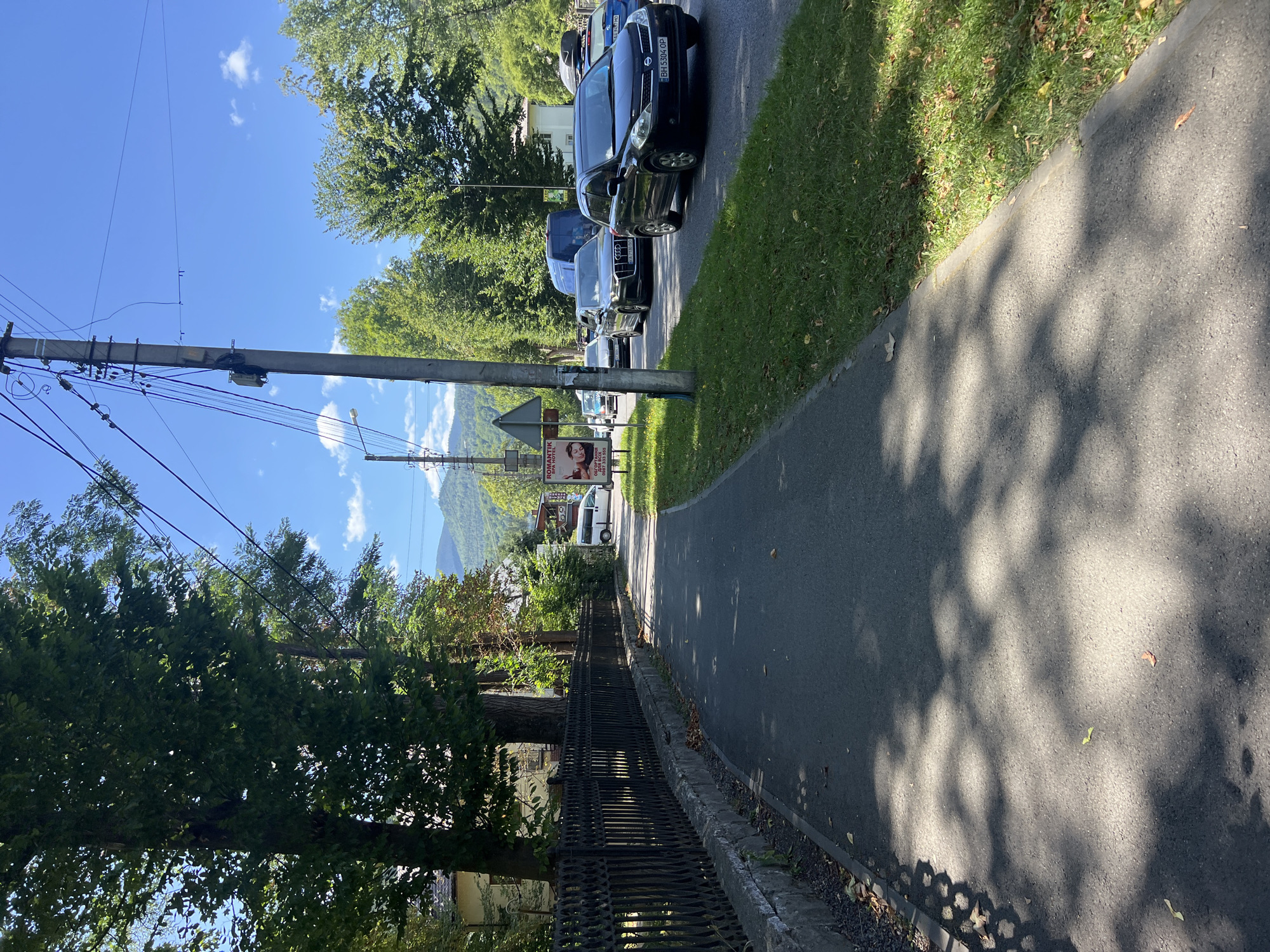 The image depicts a street scene in Yaremche, Ukraine, on a sunny day. A paved sidewalk runs along the left side of the frame, bordered by a black wrought-iron fence and mature trees. The sidewalk casts dappled shadows on its surface from the foliage above. On the right side of the street, there is a grassy verge and a line of trees.
A road runs parallel to the sidewalk, with several cars driving in the same direction. These include a black sedan, a silver Audi, a blue van, and a dark blue SUV with a roof rack. A white van is parked on the right side of the road, near a billboard. Traffic signs and utility poles with numerous wires are also visible along the roadside. In the background, lush green mountains rise against a clear blue sky with scattered white clouds. The overall impression is of a pleasant, somewhat busy, day in a town nestled within a mountainous landscape.