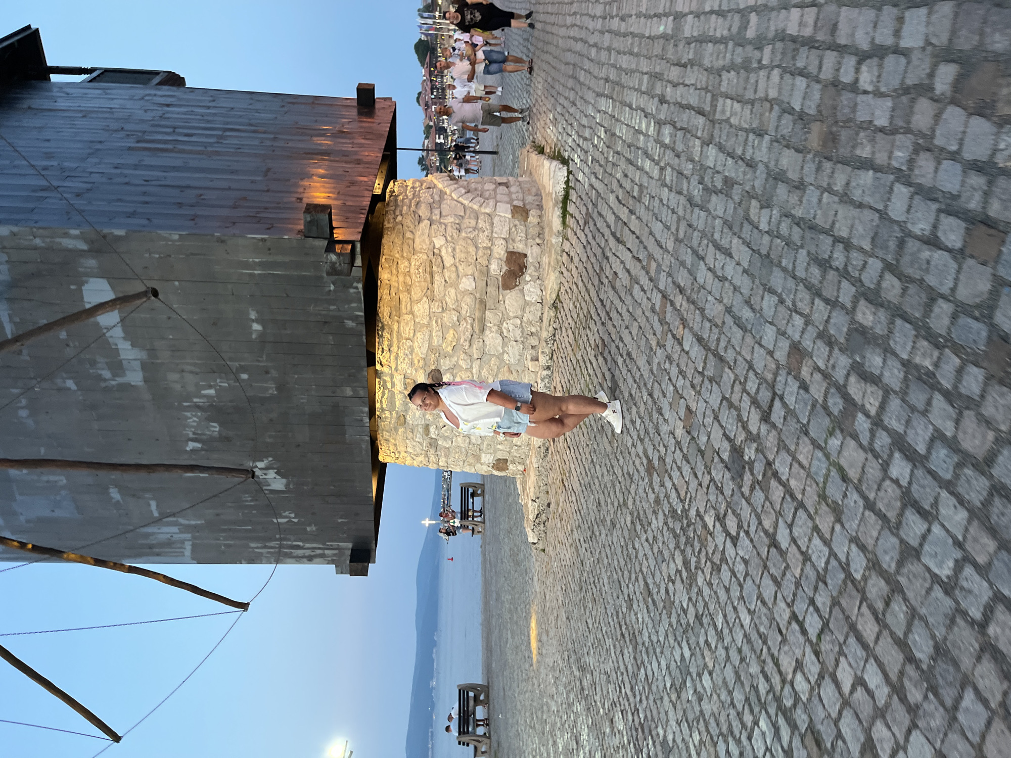 The image shows a woman standing on a cobblestone promenade in the city of Nesebar, Bulgaria, at dusk. She is wearing a white short-sleeved blouse, denim shorts, and white sneakers. She is smiling and looking towards the camera. Behind her is a large, old windmill with a stone base and a wooden tower. The windmill is illuminated by warm light, casting a glow on the stone base.
In the background, a calm sea stretches out to the horizon, with a distant coastline visible. Several people are walking along the promenade, and a few are sitting on benches near the sea. The sky is a dusky blue, indicating the end of the day. The overall atmosphere is peaceful and scenic. The cobblestone pavement suggests a historic area, characteristic of Nesebar's Old Town.
