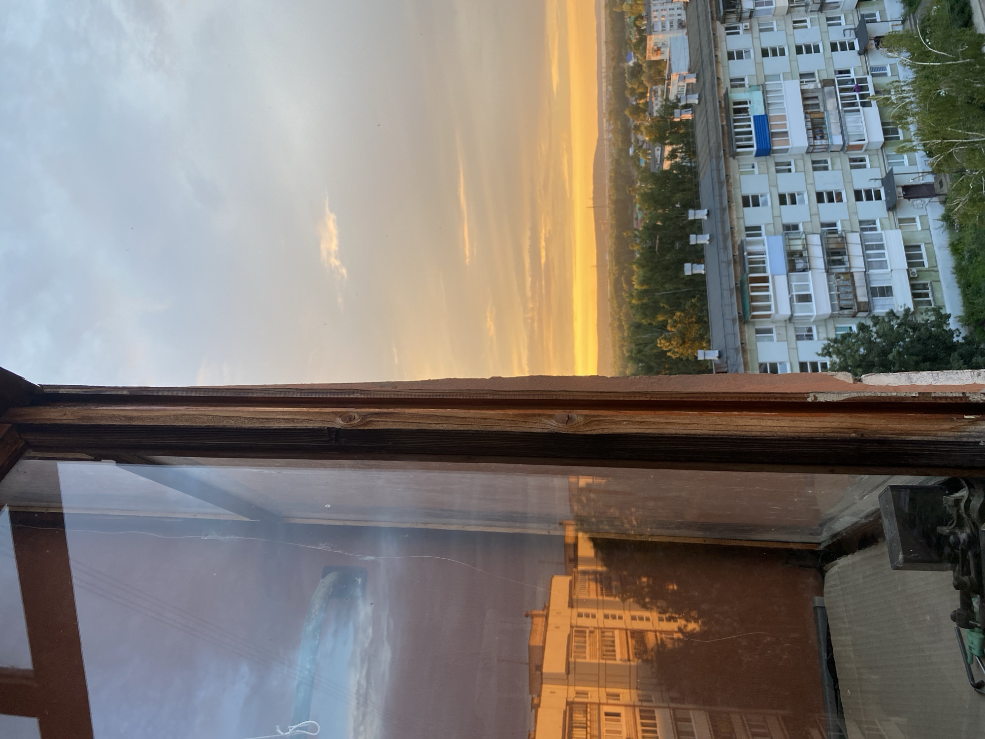 The image shows a view from a window in Chaikovsky, Russia, overlooking a residential area at sunset. The window frame is made of weathered wood, and the glass is dirty with visible cracks. A reflection in the window shows a multi-story building bathed in the warm, golden light of the setting sun.
Outside the window, a landscape of trees and buildings is visible. In the foreground, a light blue, multi-story apartment building with balconies and numerous windows dominates the view. The sky is a gradient of pale blue and orange, with wispy clouds, indicating the late afternoon or early evening. The overall scene suggests an urban setting in a residential district, likely in the outskirts of Chaikovsky, during a calm, transitional time of day. There are no people visible in the image.