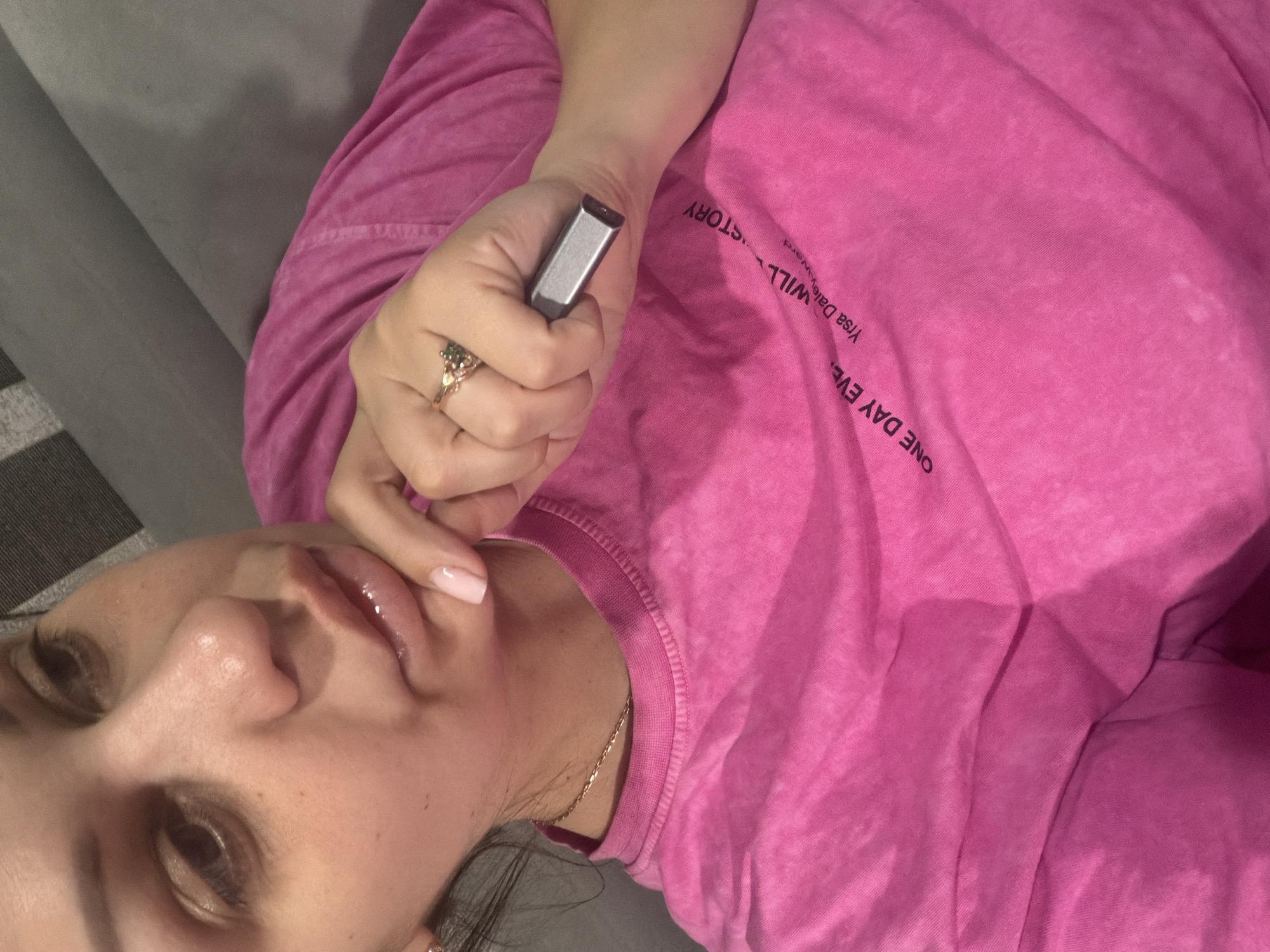 A woman is seen in a close-up shot, her head tilted to the side. She is wearing a bright pink t-shirt with black text printed on it, which appears to be a quote or a name. Her right hand is raised to her chin, with her thumb tucked under her chin and her index finger resting on her lip. She is wearing a ring on her right index finger and a delicate gold necklace around her neck. A silver electronic device, possibly a vape or a USB drive, is held in her right hand. Her fingernails are painted a light pink. The background is a soft gray, suggesting she is indoors, possibly on a couch or a chair. The lighting is dim, with some areas of the t-shirt appearing lighter due to creases and light reflection. There is no discernible indication of the image being taken in Sochi, Russia.