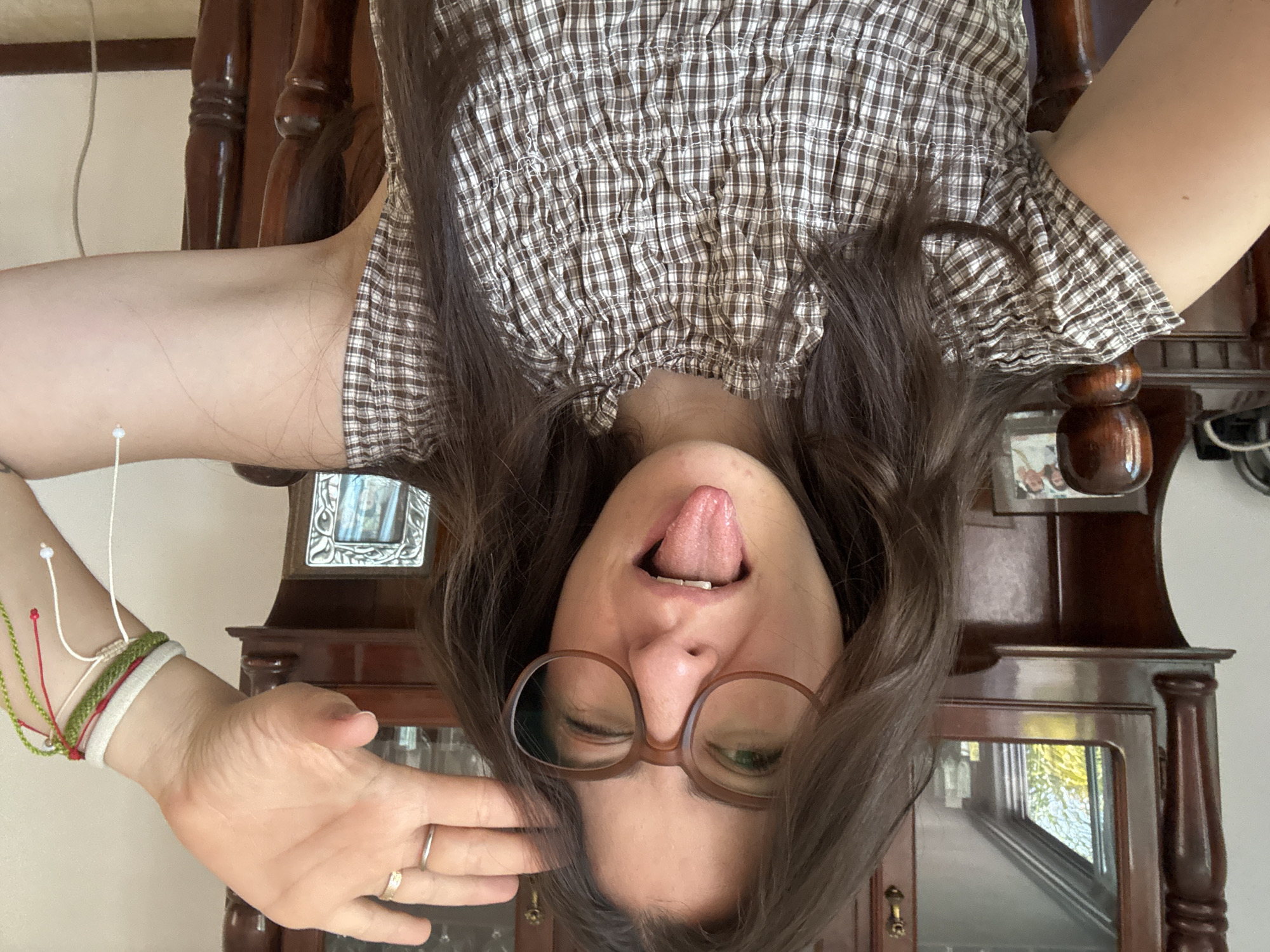 An upside-down portrait captures a person, likely a young female, making a playful face. The individual has long, dark brown hair that falls around their face due to the inverted orientation. They are wearing a brown and white checkered or plaid top with a gathered or shirred texture. Large, brown-framed glasses are worn, and the person is winking with their left eye (from the viewer's perspective) while sticking out their tongue.
Their left hand is raised, with the palm facing outwards, showing a silver ring on the ring finger and possibly a second ring. Several bracelets are visible on their wrist, including green, white, and red string-like bands, along with a white elastic band. A white string or cord is also visible near their arm, possibly belonging to an earring or accessory.
The background consists of dark wooden furniture, possibly a hutch or cabinet, featuring carved details and glass panels with reflections. A glimpse of a window or light source is reflected in the glass. A plain white wall is visible behind the furniture. The lighting suggests an indoor setting during the daytime. No visible text is present in the image. The overall mood is informal and humorous.