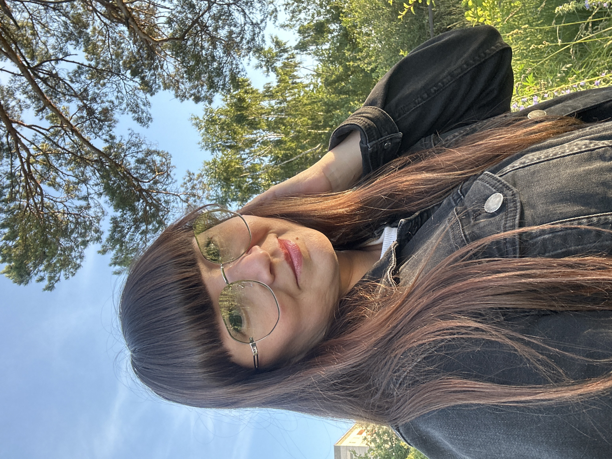 The image shows a medium shot of a woman with long brown hair and bangs, wearing glasses and a dark denim jacket. She is outdoors with trees and a building in the background, under a clear sky. The lighting suggests it is late afternoon or early evening. The woman is looking towards the camera, with her right hand touching her hair. The environment appears to be a park or natural area within the city of Chaikovsky, Russia, given the presence of trees and foliage common to the region, and the architectural style of the building visible.