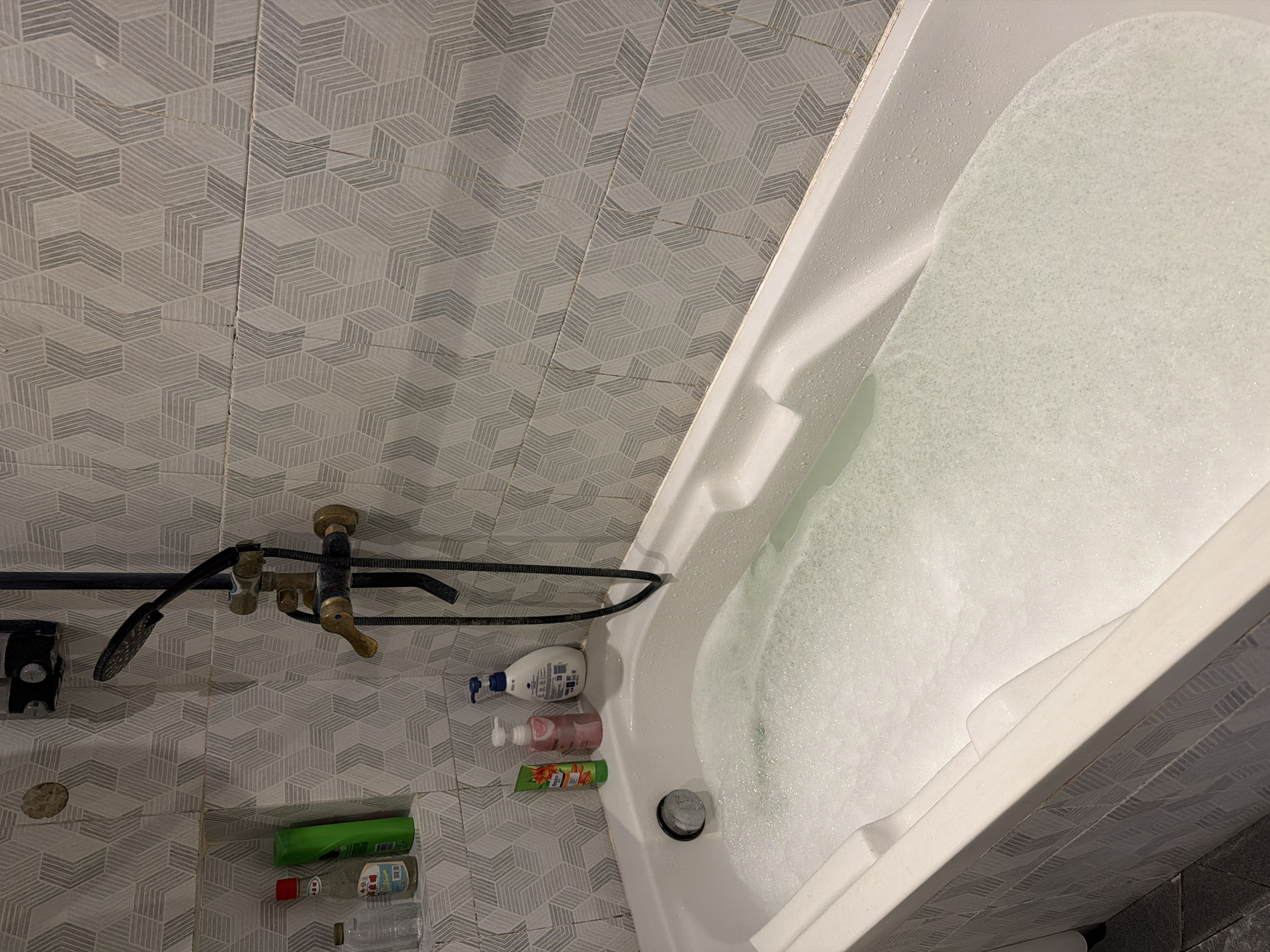 The image shows a bathroom in Denpasar Barat, Indonesia. The walls are covered in light gray geometric patterned tiles. A white bathtub is partially filled with bubbly water. A black shower fixture is mounted on the wall, along with a brass faucet. Below the fixture, several bottles of toiletries are visible, including shampoo, body wash, and other personal care products. There is a brown stain on the wall.