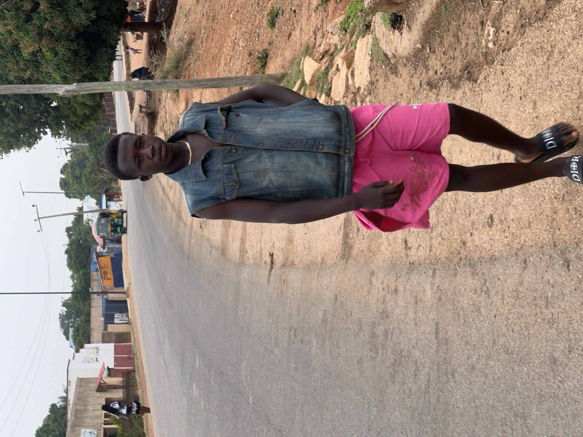 A person stands on the side of a road. They are wearing pink shorts, a blue denim vest, and black sandals with white "DIOR" logos. The person has dark skin and short hair. In the background, a road stretches into the distance, with buildings and trees visible on either side. Utility poles line the road. The setting appears to be in Du, Nigeria.