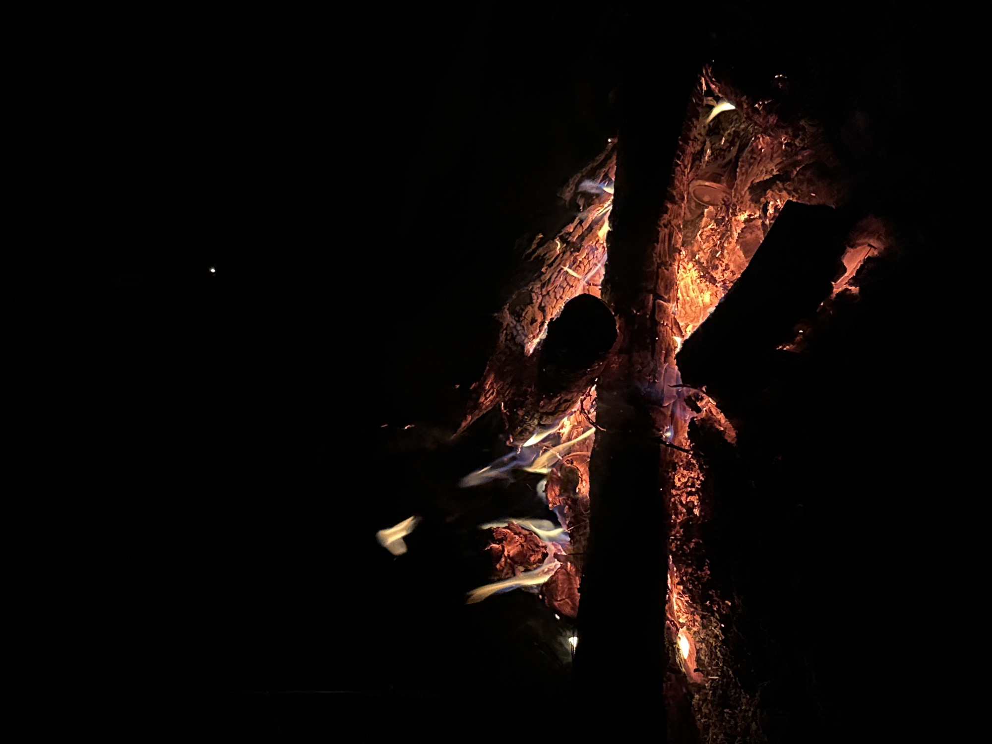 In the dark, an open campfire burns with orange and white flames around several large pieces of wood. A small white light, possibly a star, appears in the upper left corner. The location is presumably Юзуфоўскі сельскі Савет, Belarus, based on the prompt.