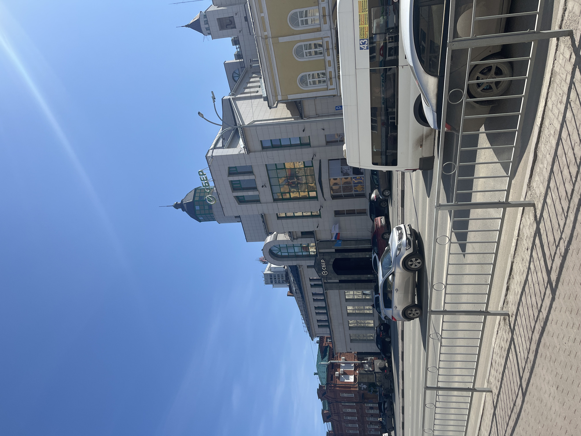 The image shows a street scene in Ulyanovsk, Russia. Several buildings line the street, including a yellow building with arched windows and a gray building with a sign that reads "Сбер" (Sberbank) in Russian. Cars and a bus are parked along the side of the street. A metal barrier stands between the sidewalk and the street. The sky is blue with few clouds.