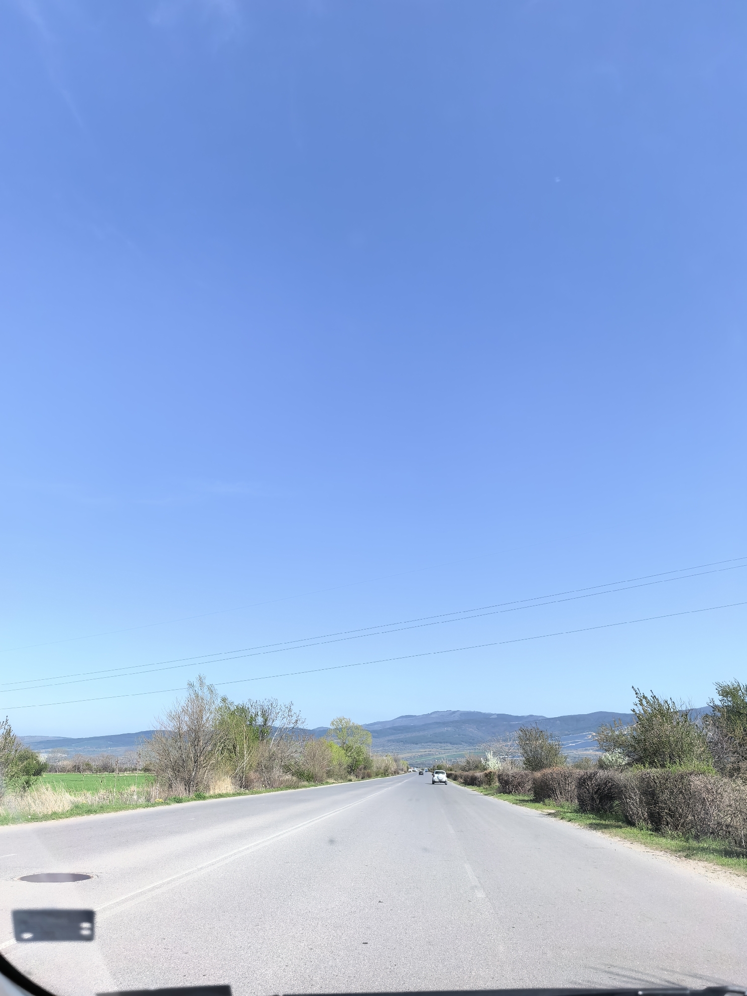 The image captures a road trip scene from the perspective of a vehicle's dashboard. A wide, two-lane asphalt road extends into the distance under a vast, clear blue sky with only faint traces of wispy clouds. The road features a white dashed line separating the lanes and a solid white line on the right shoulder. A few cars are visible far ahead on the road, with a white SUV being the most distinct.
On the left, there are green fields and scattered trees, some still bare and others with early spring foliage. The right side of the road is lined with dense bushes and more deciduous trees. In the background, distant, hazy blue mountains or hills frame the horizon. Multiple thin power lines stretch horizontally across the mid-ground.
The time of day appears to be late morning or early afternoon, indicated by the bright sunlight and minimal shadows. The weather is sunny and clear. A manhole cover is visible on the left side of the road in the foreground. The very bottom of the frame shows reflections from the vehicle's dark dashboard. There is no visible text in the image.