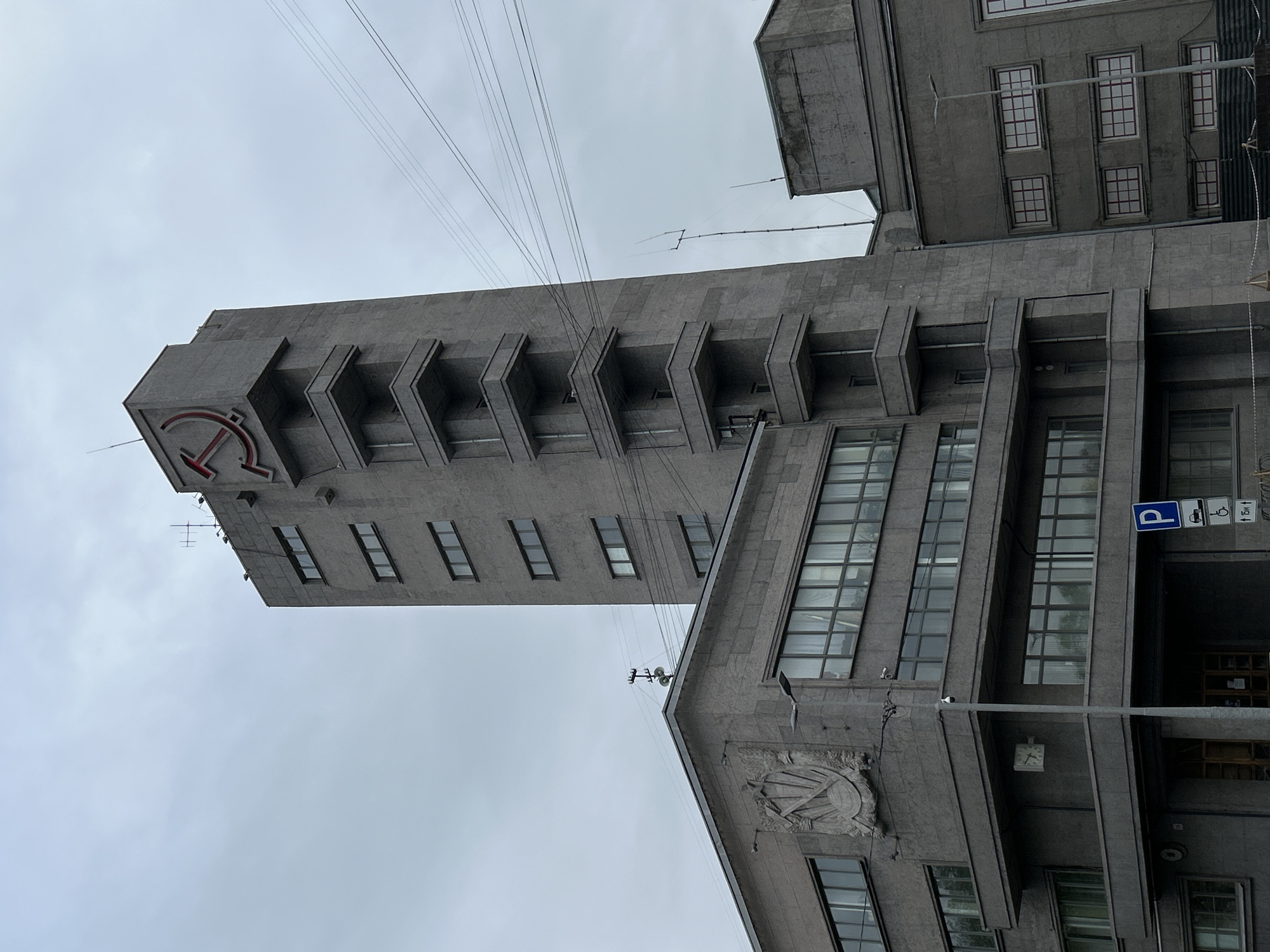 The image shows a tall, gray building in Saint Petersburg, Russia. The architectural style is Soviet, with strong vertical lines and protruding balconies. At the top of the tower is a red emblem of the hammer and sickle. Another emblem is visible on the facade of the main building. There are windows on both the tower and the main building. Street-level objects such as a parking sign and wires are visible. The sky is overcast.