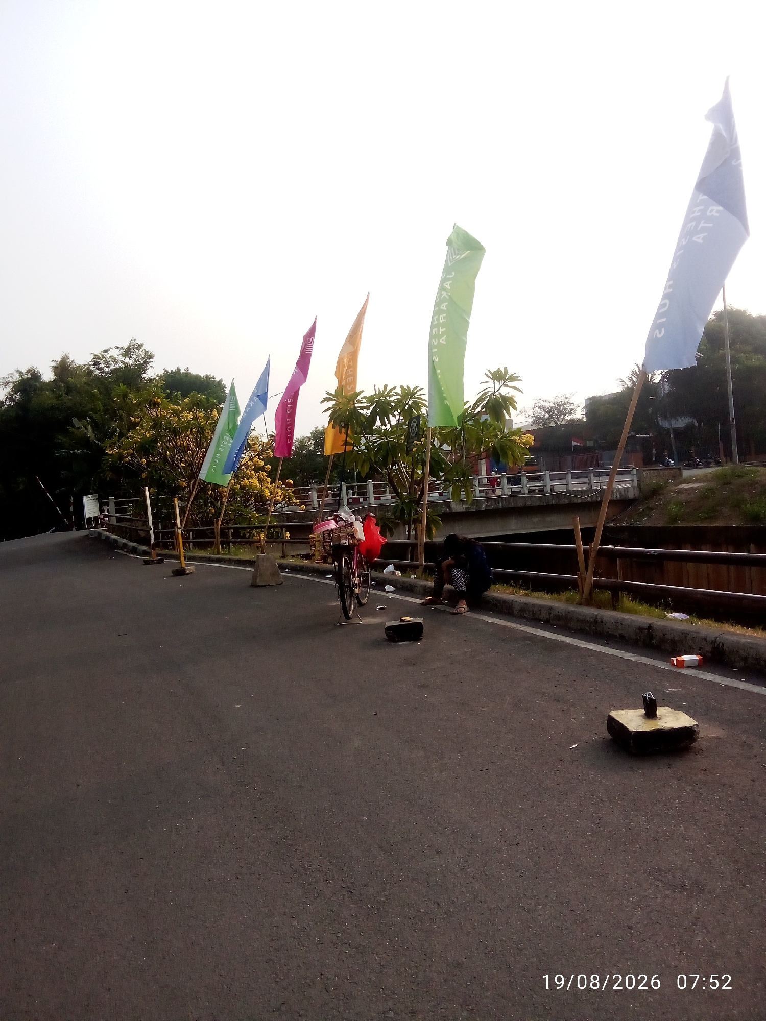 The image shows an outdoor scene with a road, a bridge over a canal, and several flags. A person is sitting on the side of the road, near a bicycle with a basket. There are trees and vegetation in the background, and the sky is overcast. The timestamp "19/08/2026 07:52" is visible in the lower right corner. The scene appears to be a street in East Jakarta, Indonesia, possibly near a waterway or park.