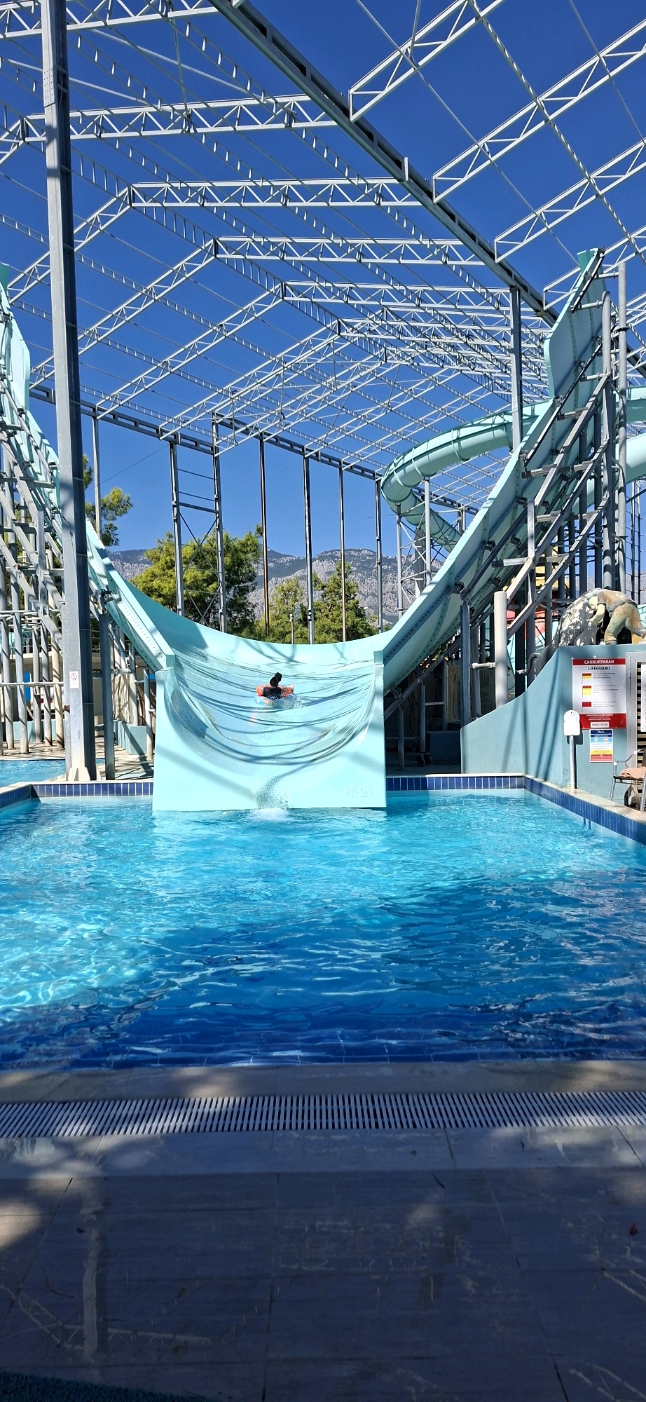 An outdoor water park in Kemer, Turkey, features a large turquoise water slide with a person on a raft descending it. The slide is framed by a metal structure and a bright blue sky. Trees and mountains are visible in the background. A swimming pool with clear blue water is in the foreground. The scene appears to be during daylight.
