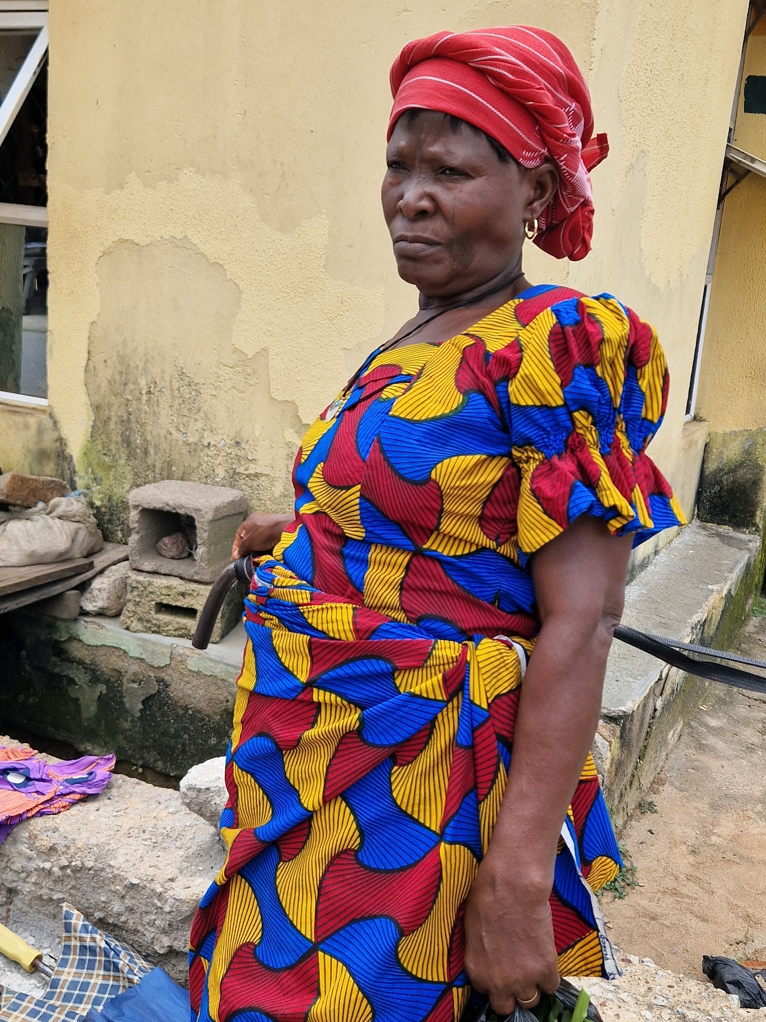 An outdoor, medium shot captures a woman with dark skin and medium build, wearing a vibrant, patterned wrap dress and a red headscarf. She has a serious expression, her gaze directed off-camera to the left. Her left arm is bent at the elbow, with her hand holding a dark plastic bag filled with green items. A gold hoop earring adorns her left ear. Behind her, a weathered, pale yellow building wall is visible, with patches of peeling paint and dark discoloration near the bottom. To the left of the woman, a white-framed window with a partially opened pane reflects the surroundings. Below the window, a collection of objects is arranged: a wooden plank, a white sack, and several stones. In the middle ground, a small, square concrete structure with an opening sits on top of another, larger concrete block. A dark, curved pipe extends from the right side of the frame, passing behind the woman. The ground in the foreground appears to be a mix of dirt and light-colored concrete. Further to the right, a black bird pecks at the ground near a pile of dark debris. The overall scene suggests an informal outdoor setting, possibly a market or a residential area in a tropical or subtropical climate.