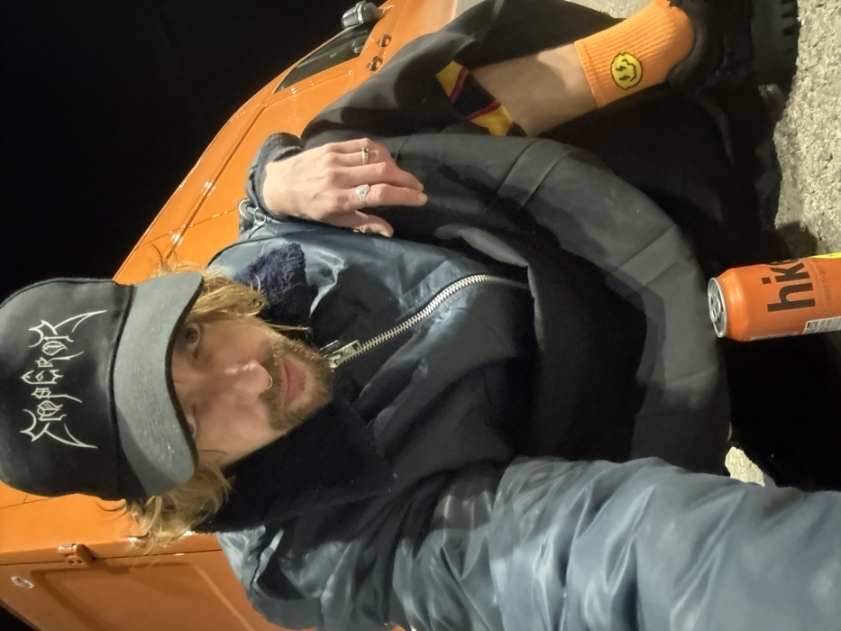 A person with long hair, a beard, and a nose ring is wearing a dark cap with white writing, a blue jacket, and an orange sock with a yellow smiley face. They are sitting or lying down on a surface next to an orange can with white writing. An orange vehicle is also visible. The person is looking at the camera.