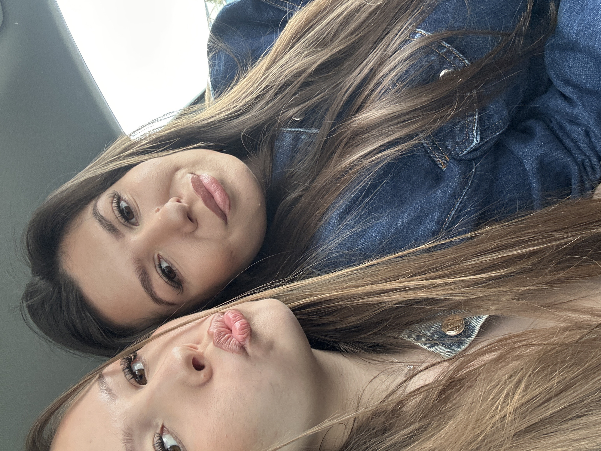 Two young women are posing for a selfie in an indoor environment, possibly inside a car based on the gray panels visible in the background. The woman on the top is looking directly at the camera with a neutral expression. The woman on the bottom is posing with puckered lips. Both have long brown hair and are wearing denim jackets. The location is not identifiable as Varna, Bulgaria, based on the visual information provided.