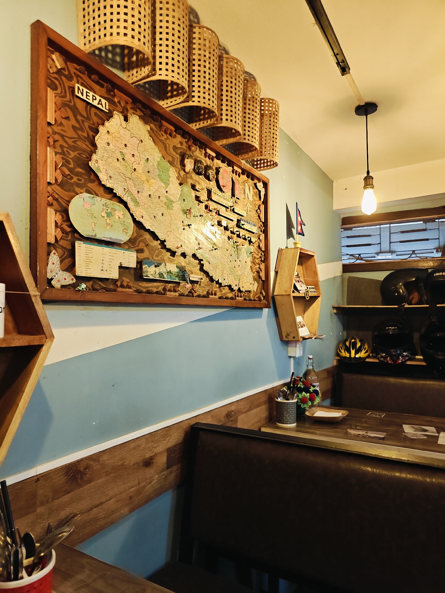 The image depicts the interior of a restaurant in Lalitpur, Nepal. A large, wooden-framed map of Nepal, adorned with pins and miniature flags, dominates the left side of the frame. Above the map, decorative bamboo lamps hang from the ceiling. To the right of the map, a small, hexagonal wooden shelf displays books and other items. The walls are painted in a light blue and white pattern, with a wooden wainscoting along the bottom. In the foreground, a dark brown booth seating area is visible, with a wooden table laid out with cutlery in a red container, a bottle of water, and a small dish. On shelves behind the booth, several motorcycle helmets are displayed, suggesting the establishment might cater to motorcycle enthusiasts or be located in an area with a significant motorcycle culture. The lighting is warm and ambient, contributing to a cozy atmosphere. No people are visible in the frame.