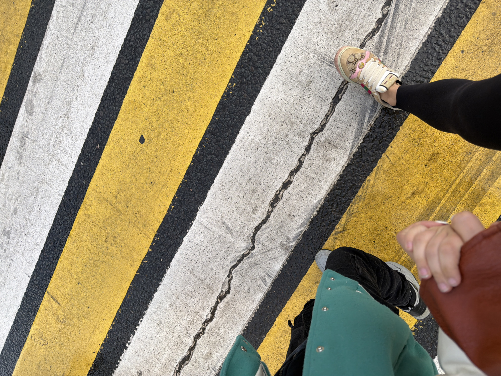 The image is a top-down view of a pedestrian crosswalk with alternating white, black, and yellow stripes. A person wearing black leggings and a tan-colored sneaker with pink accents is positioned on one of the white stripes. Another person, wearing dark jeans, a gray sneaker, and a turquoise jacket, is visible near the bottom right of the frame, holding hands with another person who is wearing a brown jacket. The crosswalk appears to be cracked. The location is unknown.