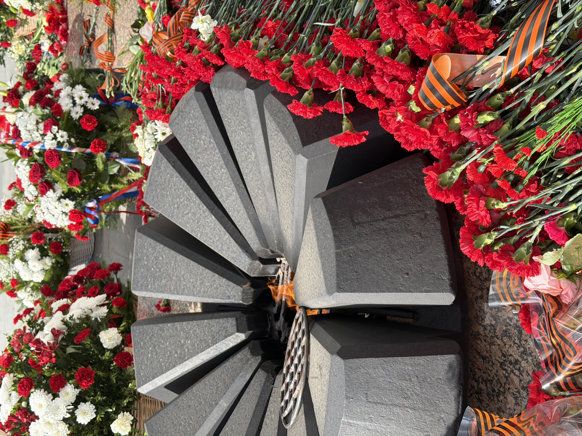 The image shows an Eternal Flame memorial in Chaikovsky, Russia, surrounded by flowers. The memorial is constructed of dark gray stone blocks arranged in a circular pattern with a fire burning inside. Wreaths and bouquets of red and white flowers are placed around the memorial, adorned with ribbons of orange and black stripes.