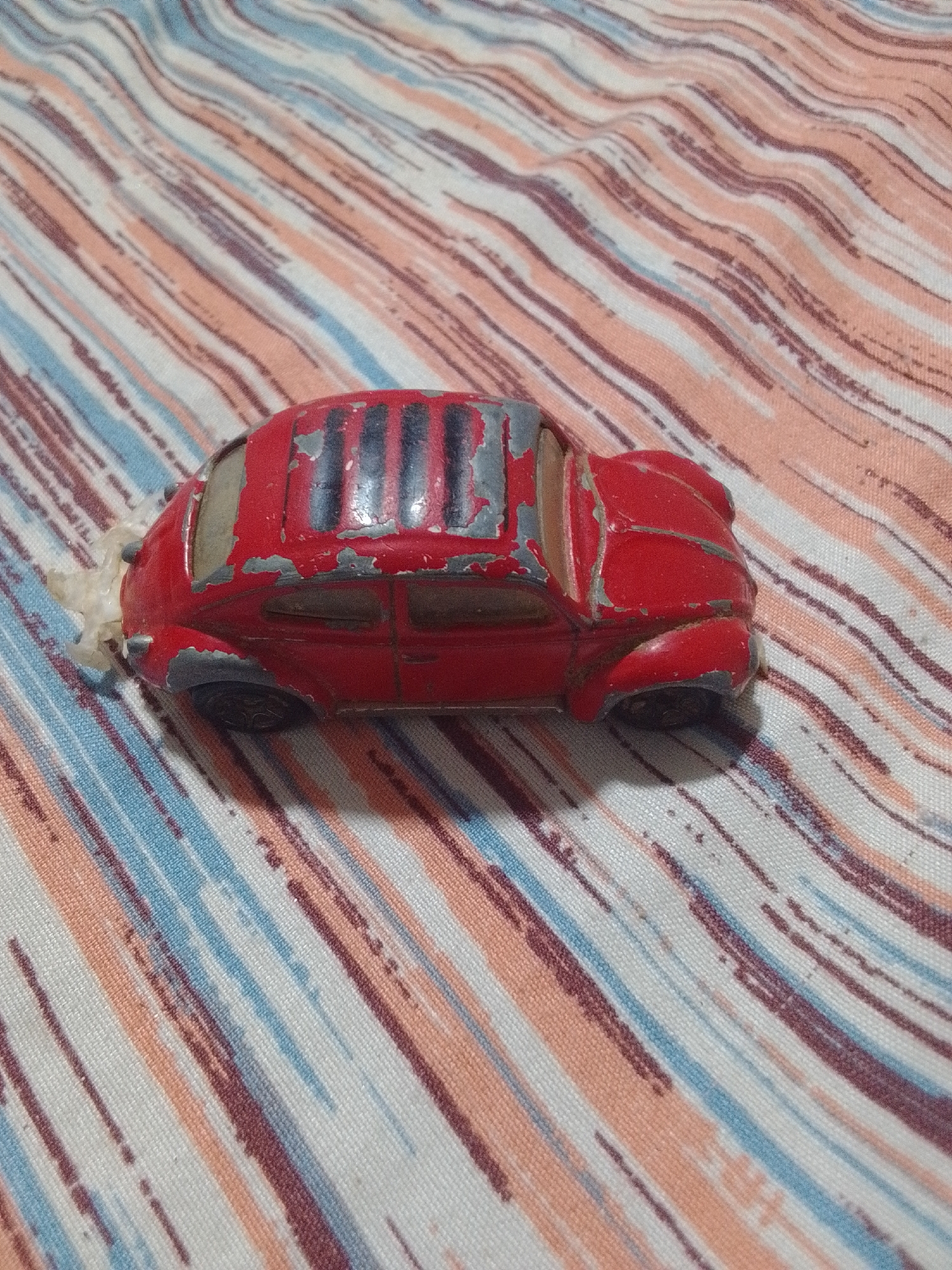 A red toy car, resembling a Volkswagen Beetle, sits on a striped fabric surface. The car shows signs of wear, with paint chipped off in several areas, revealing a grey undercoat. It has black stripes on its roof and black wheels. A white, fluffy substance is attached to the rear left side of the car. The fabric beneath has a pattern of diagonal stripes in various shades of pink, blue, and white. There is no indication of the location being the city of La Tagua, Venezuela.