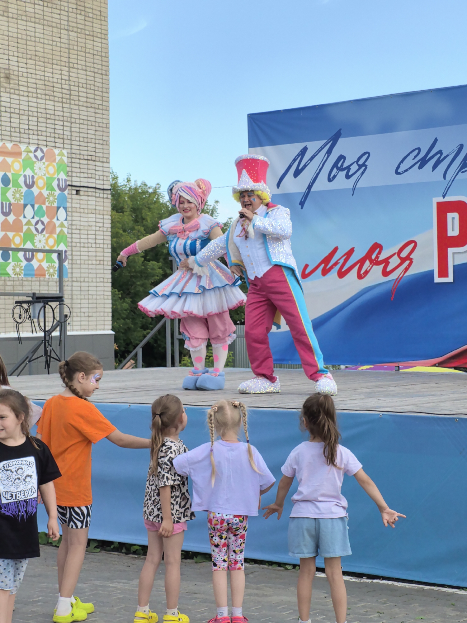 The image shows an outdoor event in the city of Фролы, Russia. A stage is set up with a blue backdrop featuring white stripes and Russian text. Two performers, dressed in colorful costumes, are on the stage. One performer, wearing a pink and blue layered dress and a colorful headdress, appears to be singing into a microphone. The other performer, in a sparkling white jacket, pink pants, and a tall red hat, is also singing and gesturing. In the foreground, several young girls are standing on the ground, watching the performance. They are casually dressed, with some wearing t-shirts and shorts. Behind the stage, a brick building and trees are visible, suggesting an urban or suburban setting. The overall atmosphere appears festive and family-friendly.