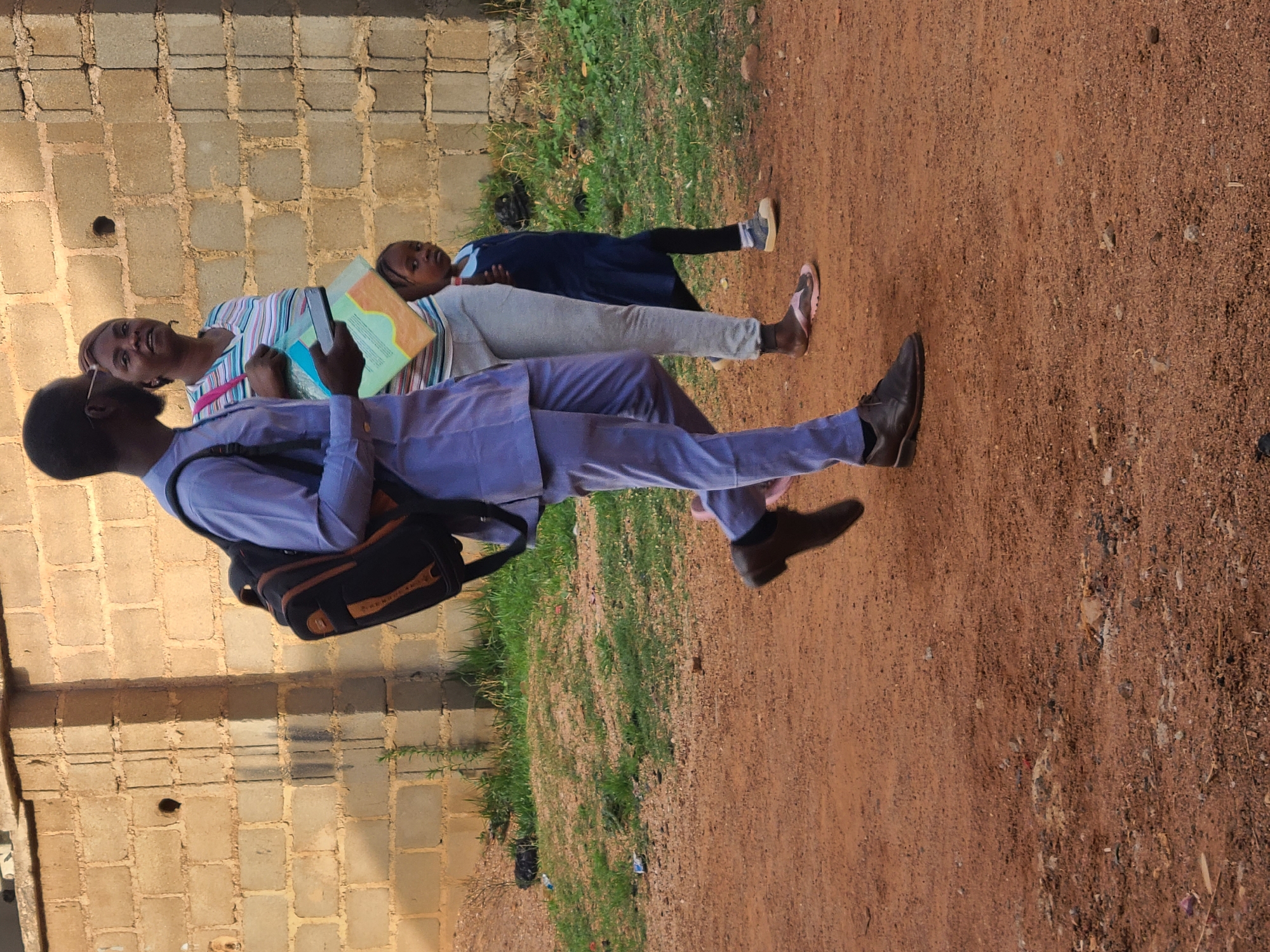 The image depicts three individuals in an outdoor setting in Barakin Lerawi, Nigeria. A man in a light purple suit and black shoes walks toward the right of the image, carrying a black backpack. A woman in a striped shirt and light gray pants walks alongside him, holding what appears to be a textbook or notebook. A young girl in a navy blue dress and light gray leggings walks next to the woman. The ground is a mixture of red dirt and green grass. To the left is a wall constructed of light brown bricks.