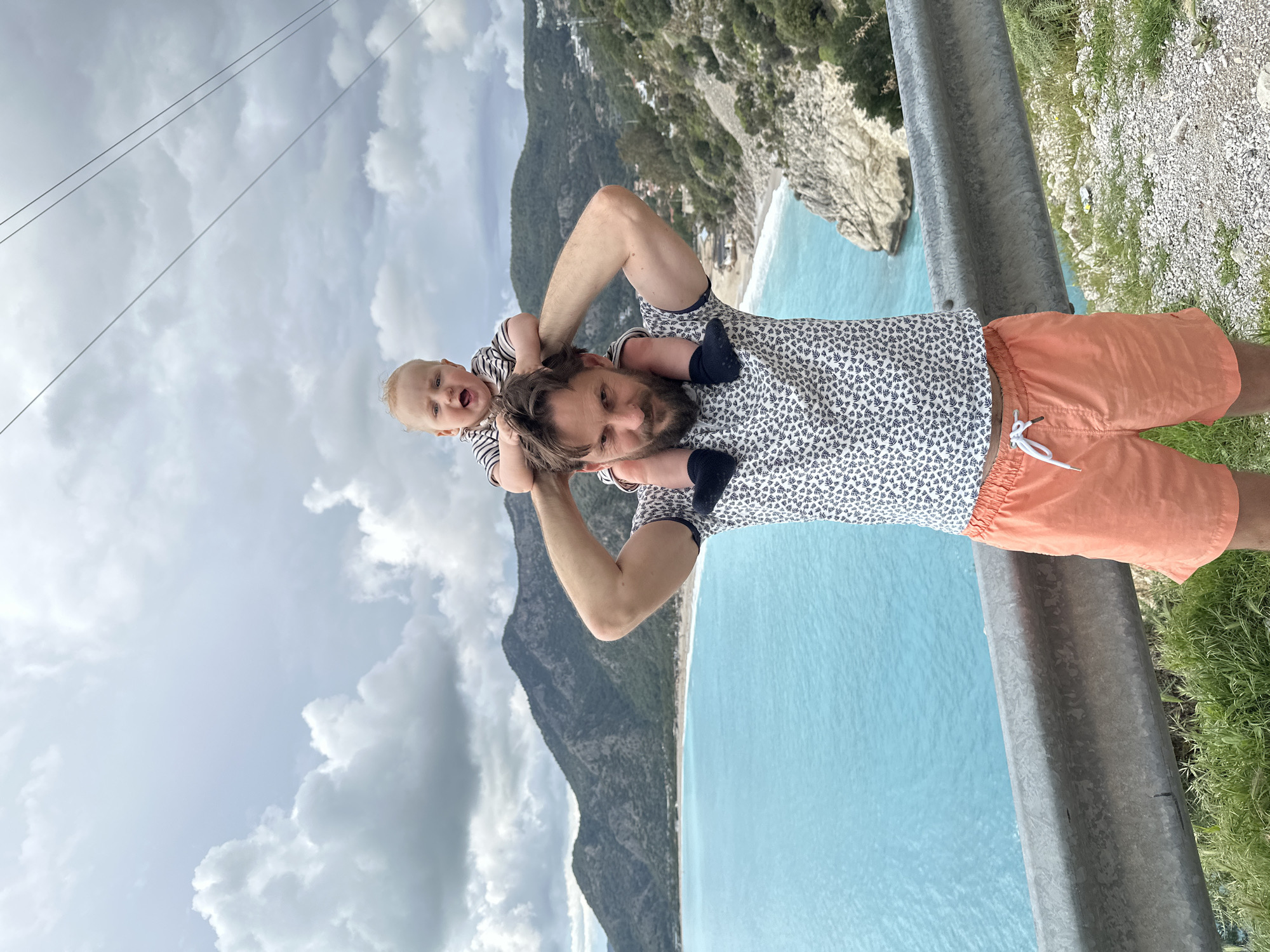 A man with a beard stands beside a metal guardrail with a baby on his shoulders. The man wears a printed short-sleeved shirt and orange shorts. The baby wears a black and white striped shirt. The background reveals a view of the beach and the ocean from Kaputaş Plajı, a small beach located in Fethiye, Turkey. A large mountain is partially visible to the left of the man. Overcast skies are overhead.