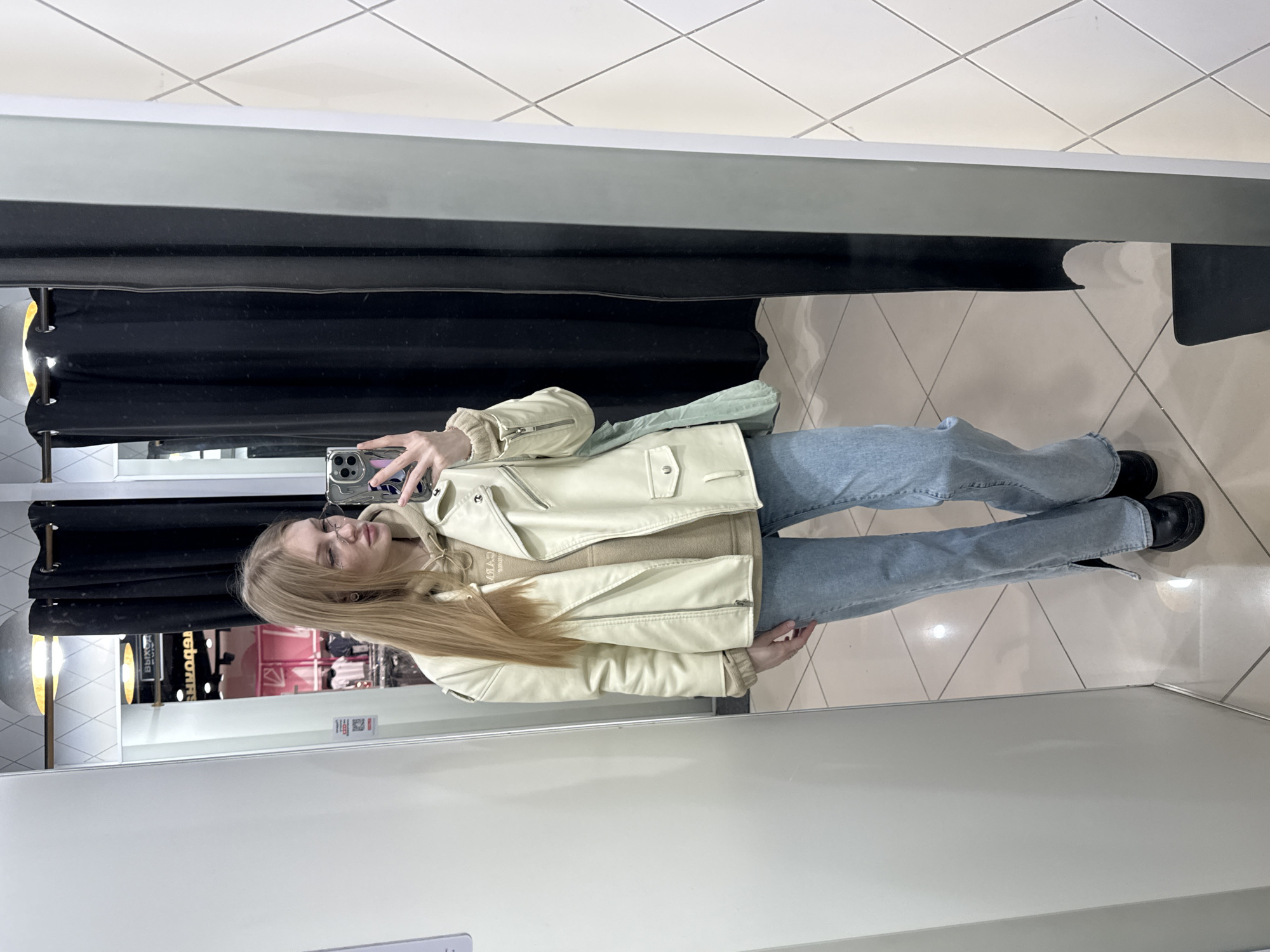 A young woman is captured in a mirror selfie, standing in what appears to be a fitting room within a retail store in Perm, Russia. She has long, straight blonde hair and is wearing glasses.
The woman is dressed in a light cream or off-white biker-style jacket, layered over a beige turtleneck or hooded sweater. She is wearing light blue, slightly flared jeans, and black shoes, possibly boots. A light blue fabric bag or garment is visible hanging from her shoulder, tucked between her arm and torso. Her right hand holds a smartphone, likely an iPhone given its distinctive multi-lens camera, which she is using to take the photograph in the mirror.
The setting is an indoor space with light-colored walls and a floor covered in square, light-colored tiles laid diagonally. Behind the woman, a black curtain typical of a changing room is visible. To the left, a silver-colored circular light fixture is present.
The reflection in the mirror offers glimpses of the store environment outside the fitting room, showing clothing racks in the background with garments and a sign. Visible text in Cyrillic, "ВЫХОД" (VYKHOD), meaning "EXIT," confirms a Russian-speaking location, aligning with the provided context of Perm, Russia. A QR code is also discernable on a sign in the reflection. The woman's expression is neutral, suggesting she is posing for the photo. The time of day is not discernible as the scene is indoors.