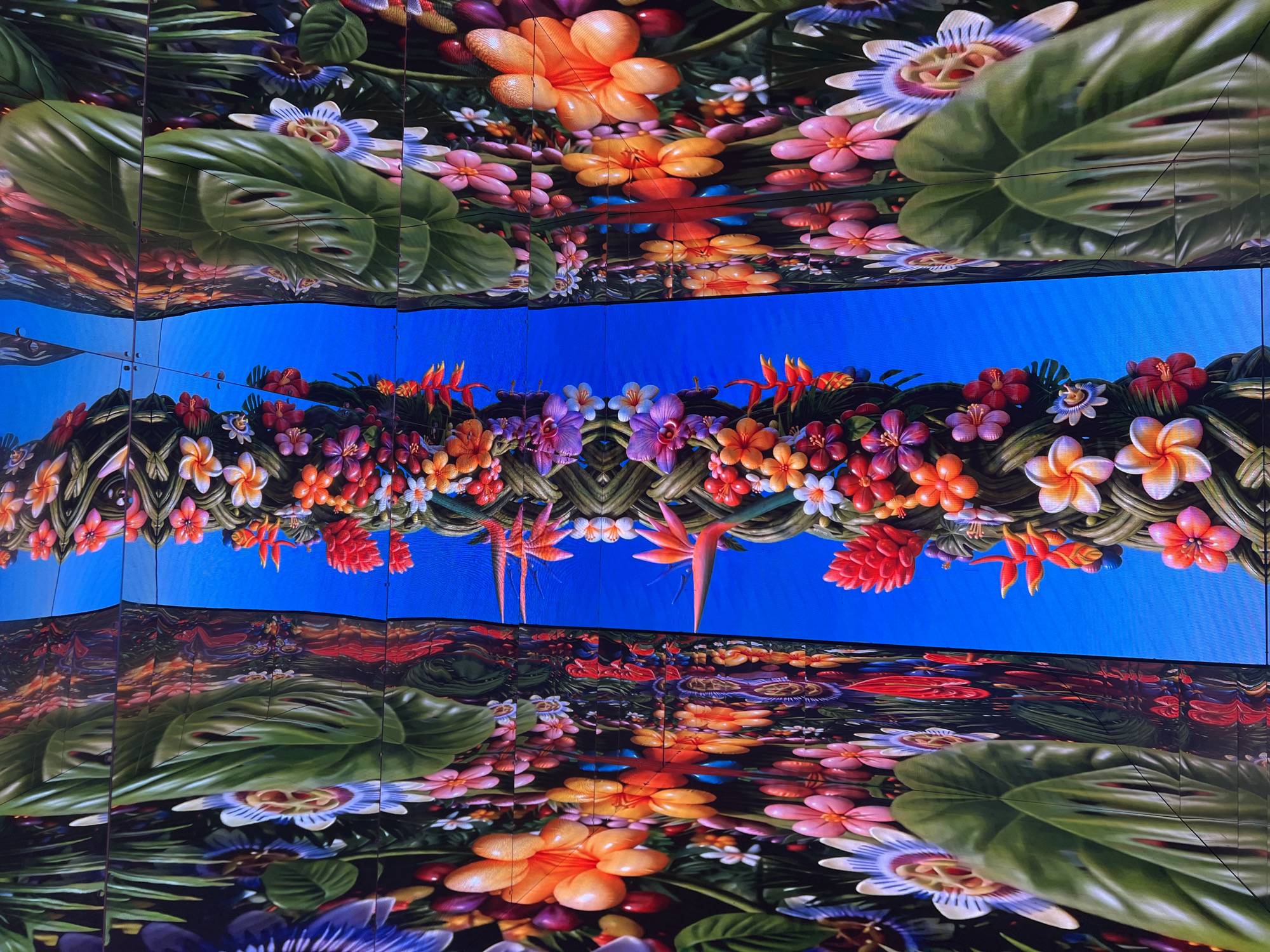 The image shows a brightly lit, digitally rendered display of lush tropical foliage and vibrant flowers, set against a clear blue sky. The artwork is presented as a diptych, with the central vertical element appearing as a twisted vine or column adorned with a cascade of colorful blooms in shades of orange, pink, purple, red, and white, resembling plumeria, hibiscus, and passionflowers. Large green leaves, possibly from a Monstera plant, frame the central composition on both sides, creating a sense of depth and abundance. The overall effect is surreal and dreamlike, with the reflective surface of the display creating a mirrored or warped effect, particularly in the blue sky background. This artwork is part of a public art installation in Miami, Florida, likely a digital mural or a screen display in a prominent outdoor location such as a plaza or a building facade. The vibrant colors and tropical theme are characteristic of Miami's aesthetic, suggesting a connection to its tropical climate and lively urban culture.