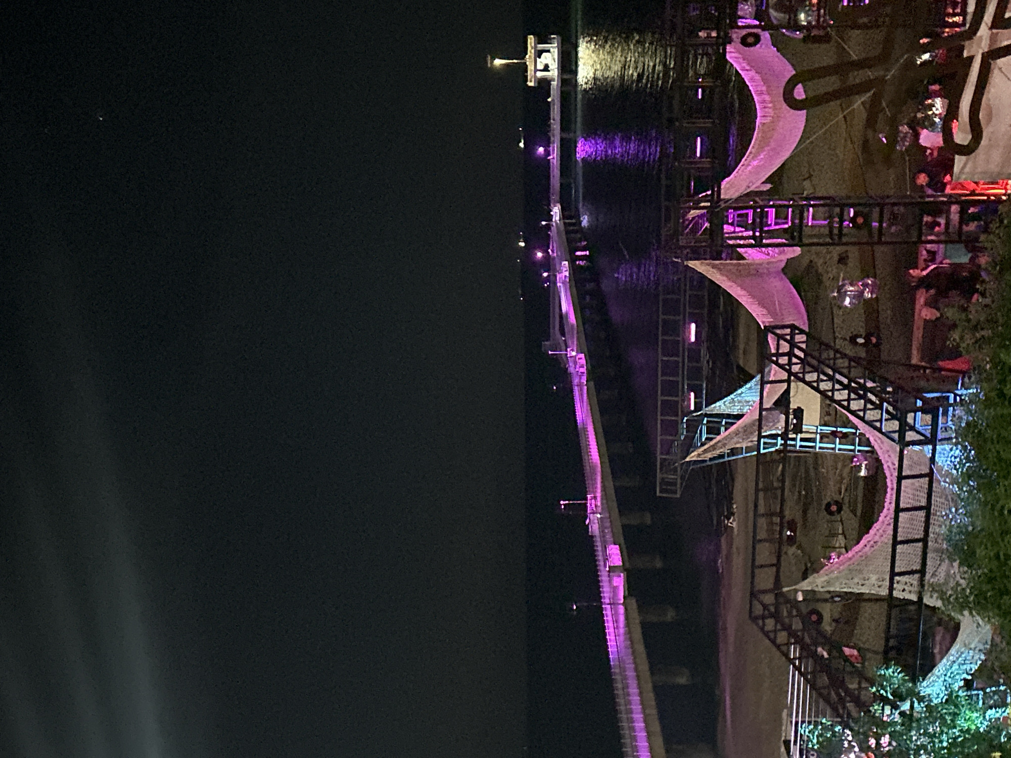 The image depicts a nighttime scene at the Burgas Pier in Burgas, Bulgaria. A pier stretches into the dark water, illuminated by purple lighting. Ornate, illuminated structures resembling nets or sails are erected on the pier. People are visible within the structure, suggesting it's a venue or attraction. The background sky is dark.