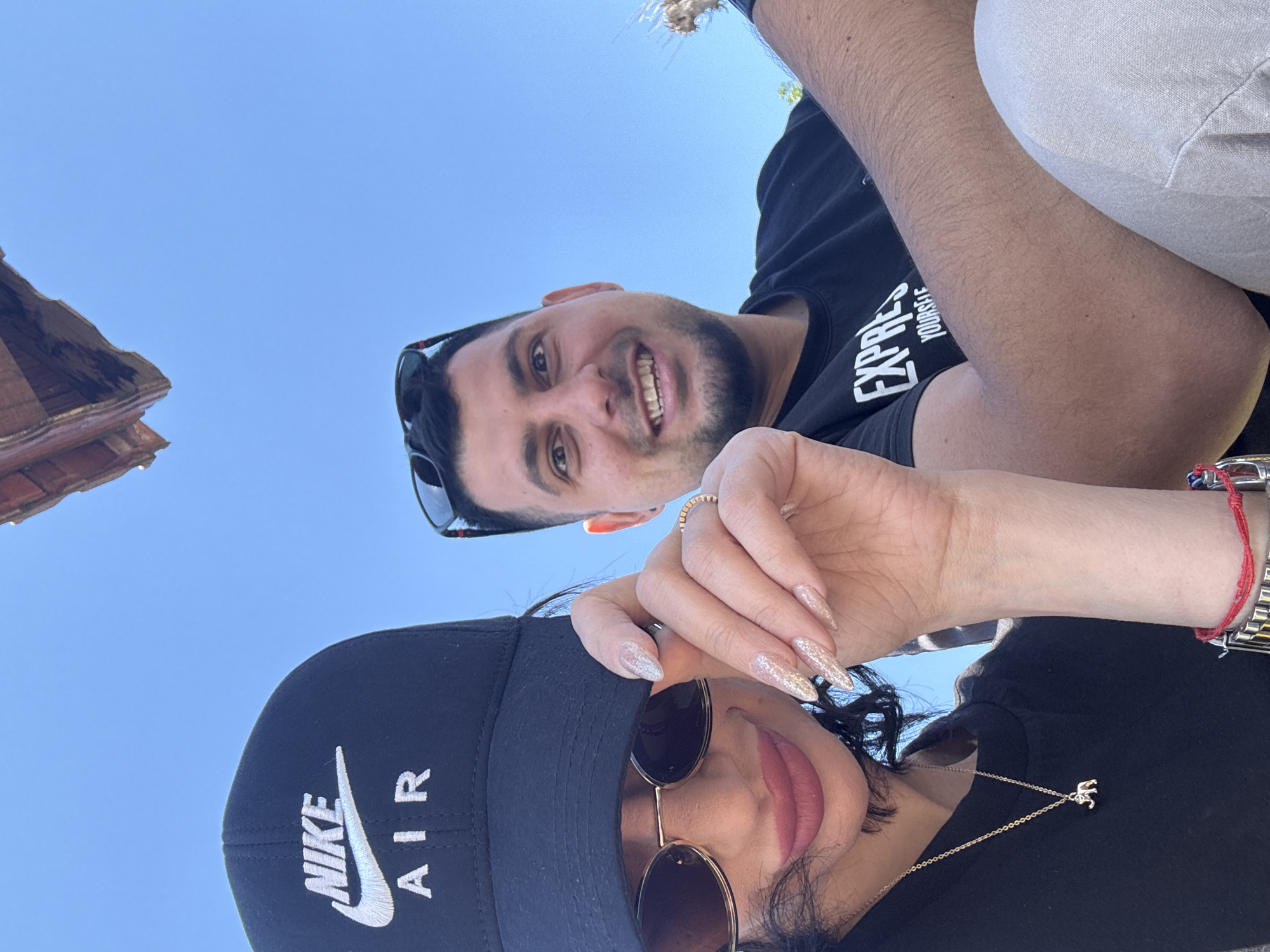 A low-angle selfie shows a man and a woman against a blue sky. A portion of a building's roof is visible in the upper left. The man, in a black T-shirt, is smiling, and his face is partially shaded by sunglasses resting on top of his head. The woman is also wearing sunglasses and a black baseball cap with "NIKE AIR" in white lettering. She has long, glittery fingernails. A gold elephant pendant hangs from her necklace. A silver wristwatch with a red string bracelet is visible on her wrist. The location is Probuda, Bulgaria, suggested by the type of architecture visible.