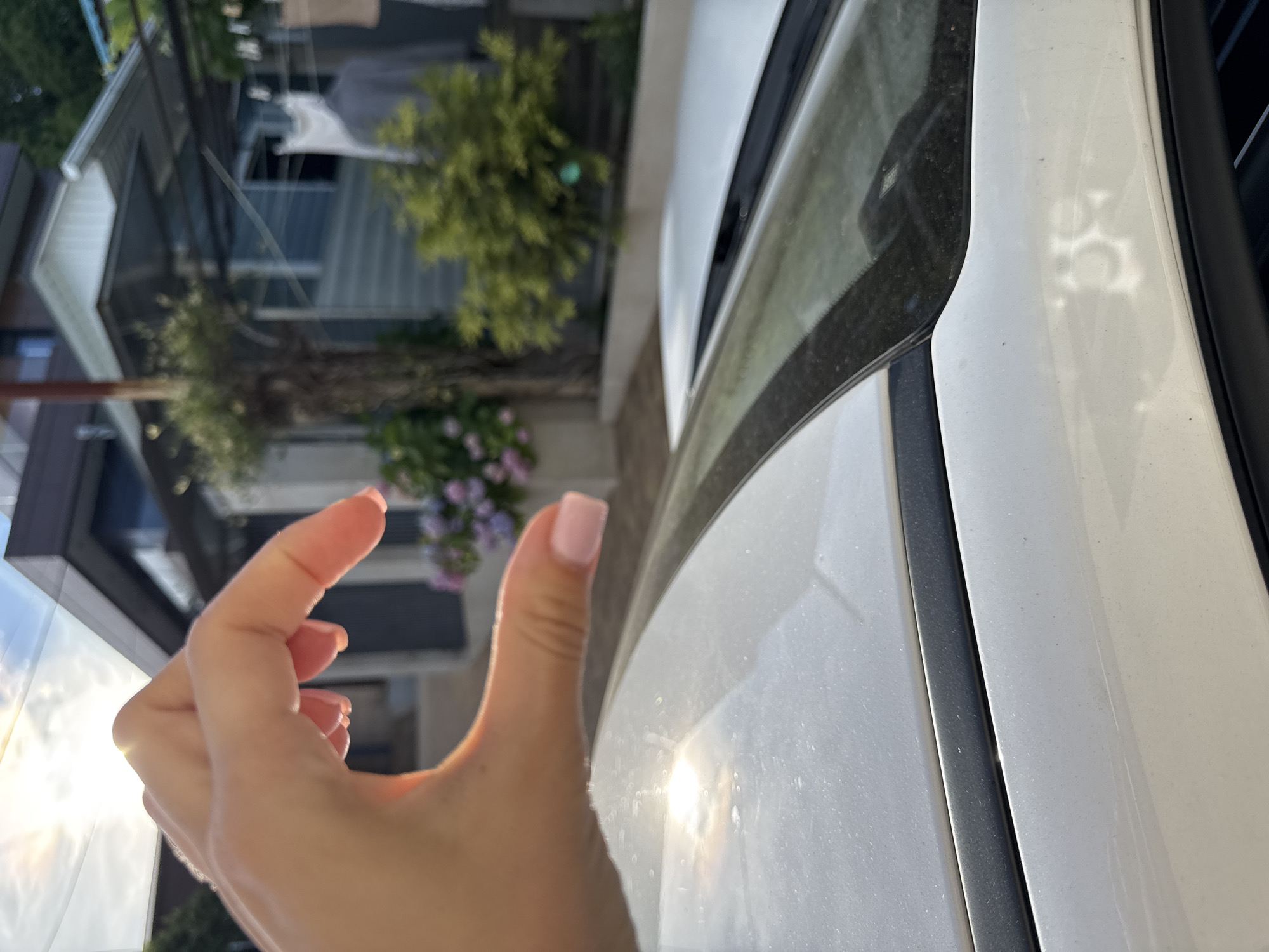 This image shows a close-up of a person's hand with painted nails, making a "pinch" gesture. The hand is positioned in front of a white vehicle, with its side and a portion of the windshield visible. The background is blurred but suggests an outdoor setting with some buildings and greenery, possibly in a residential area. The lighting indicates it is daytime, with the sun casting reflections on the car's surface. The overall mood is casual and possibly playful, given the hand gesture.
The setting is identified as Sochi, Russia. The image composition is somewhat abstract due to the close-up of the hand and the framing of the car. The focus is on the tactile gesture and the texture of the vehicle's surface.