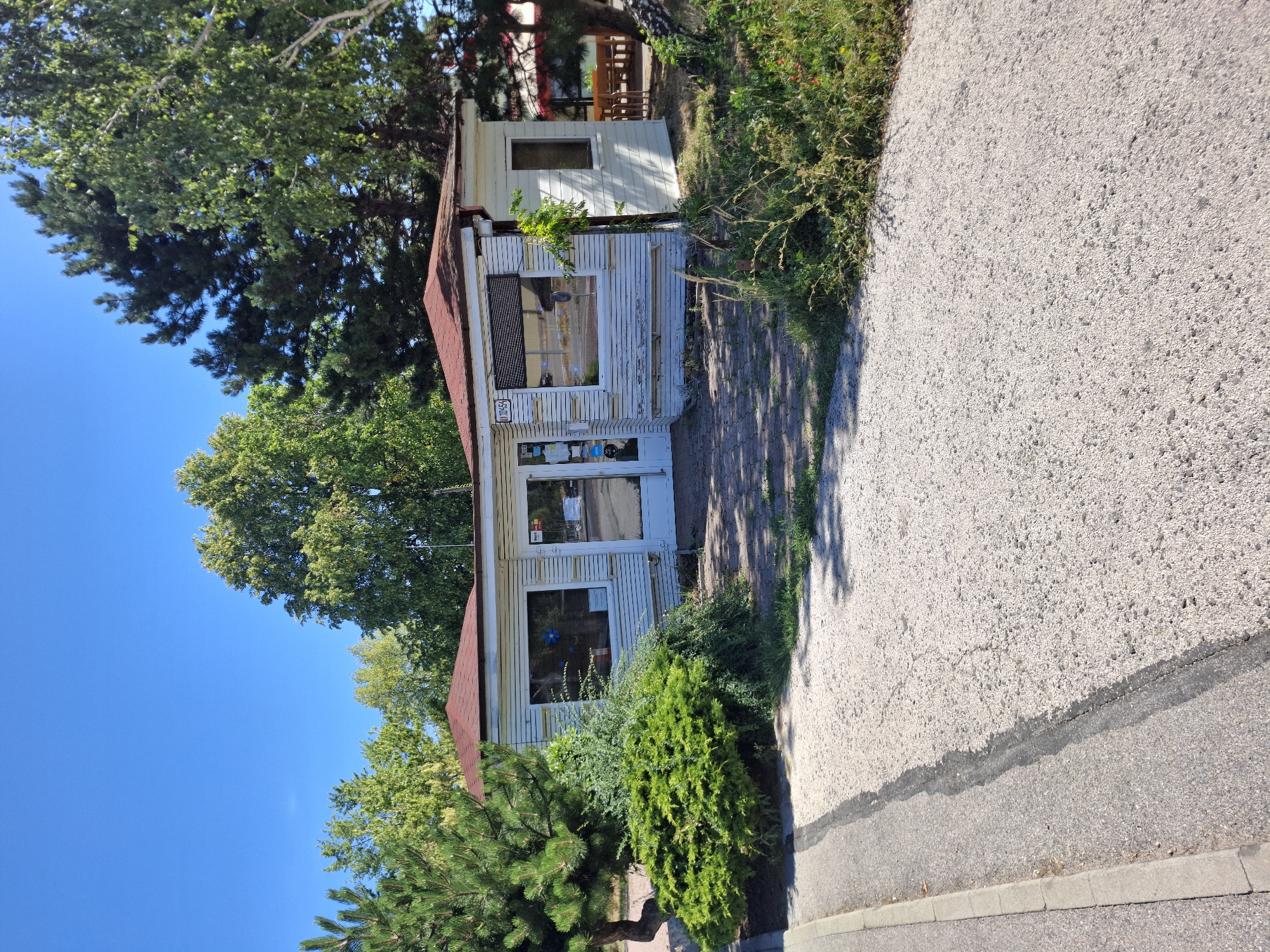 The image depicts a small, single-story building with a red, gabled roof, situated outdoors under a clear blue sky. The building appears to be an older, possibly disused structure, with white, horizontal siding that shows signs of wear and peeling paint. It has large windows and a glass-fronted door. In front of the building, there is a path paved with interlocking bricks, leading to a more general gravel-covered area. Lush green bushes and trees surround the building, with a large pine tree prominent to the left. In the background, beyond the building, a shaded outdoor seating area with wooden tables and chairs is visible, suggesting it might be a former or current cafe or restaurant. The scene has a general outdoor ambiance, likely in a park or recreational area, rather than a dense urban setting. The building's architecture and setting do not immediately suggest a specific location in Bratislava, Slovakia, without further contextual information.