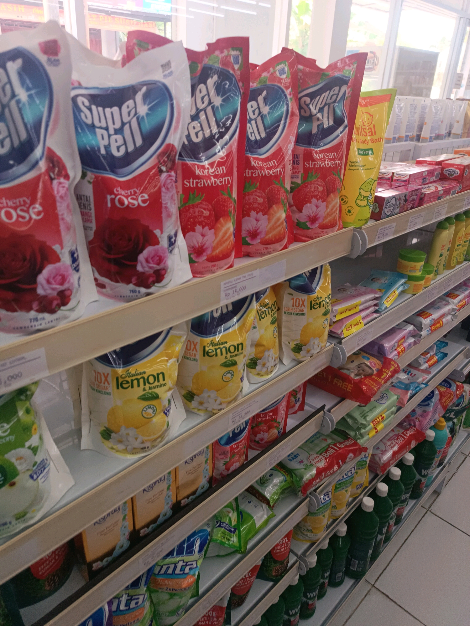 The image shows a retail shelf display in a store, likely a minimarket or supermarket, in Tabanan, Indonesia. The shelves are stocked with a variety of household cleaning and personal care products. Prominently featured are several pouches of "Super Pell" brand floor cleaner, in scents like "Cherry Rose" and "Korean Strawberry." Adjacent to these are pouches of "Vitrisal" brand hair and body bath, and other various packaged goods including laundry detergents, diapers, and bottled liquids. The products are neatly arranged on white shelves, with pricing labels visible in some instances. The lighting suggests an indoor environment, possibly illuminated by overhead store lights and natural light filtering in from a window or entrance, which is visible in the background. The overall scene depicts a typical retail merchandising environment.