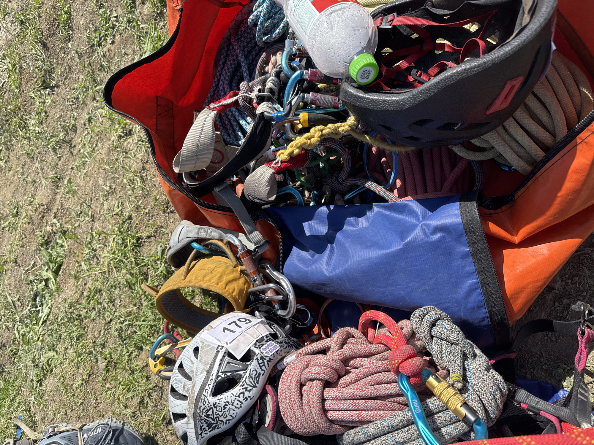The image shows a collection of climbing gear scattered on a grassy surface. Predominantly visible are ropes of various colors (red, white, gray, blue), carabiners, helmets (one black and one white with a "179" sticker and geometric designs), harnesses, and a water bottle. The gear is partially contained within an orange and blue duffel bag. The location, Khvalynsk, Russia, is not evident from the visual details of the image.