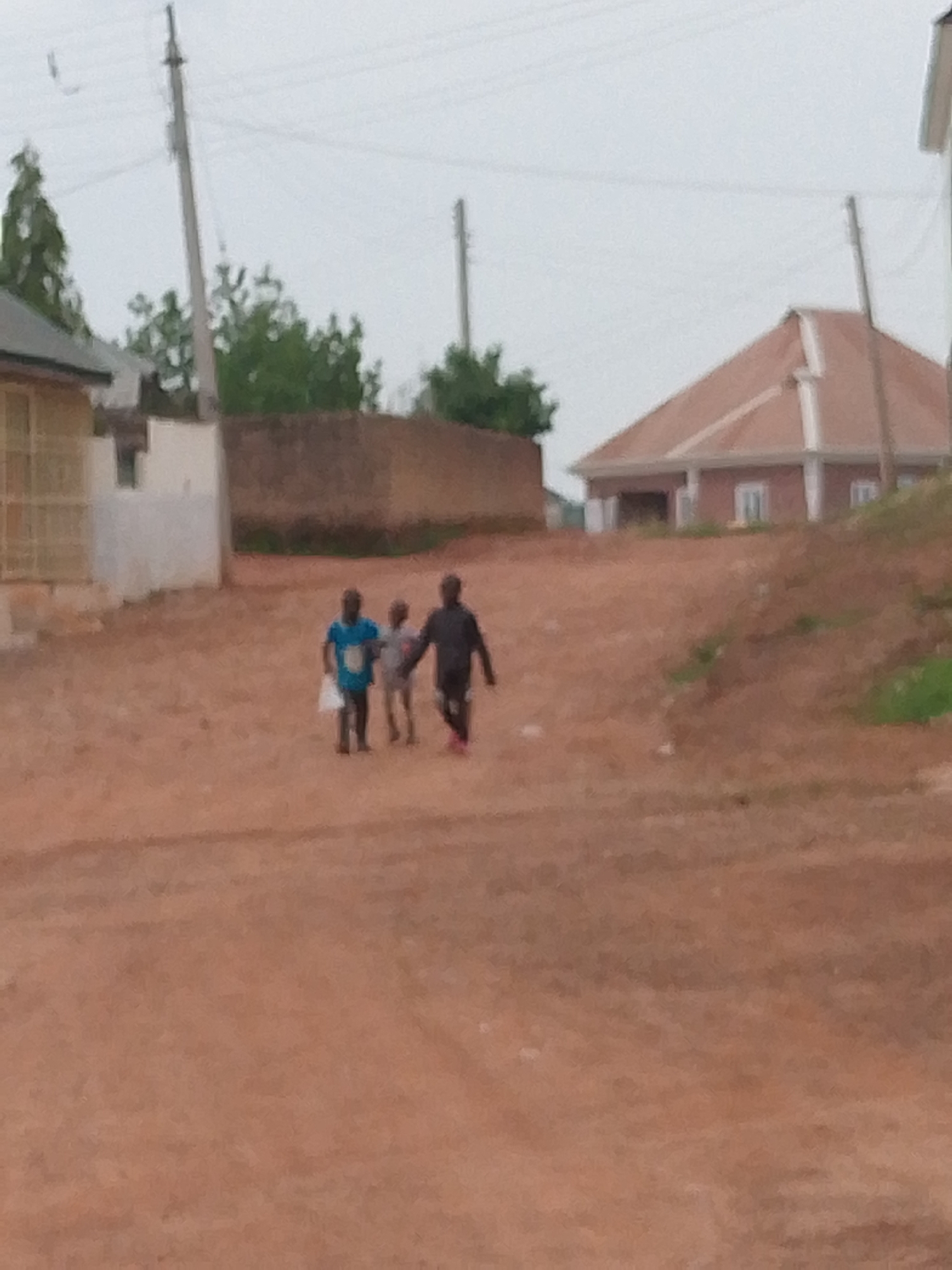 The image shows a dirt road in what appears to be a residential area in Barakin Angundi, Nigeria. Three children are walking along the road. The child on the left is wearing a blue shirt and carrying a white bag. The child in the middle is wearing a light-colored shirt, and the child on the right is wearing a dark jacket and red shoes. Buildings and vegetation are visible in the background. There are telephone poles. The sky is overcast.