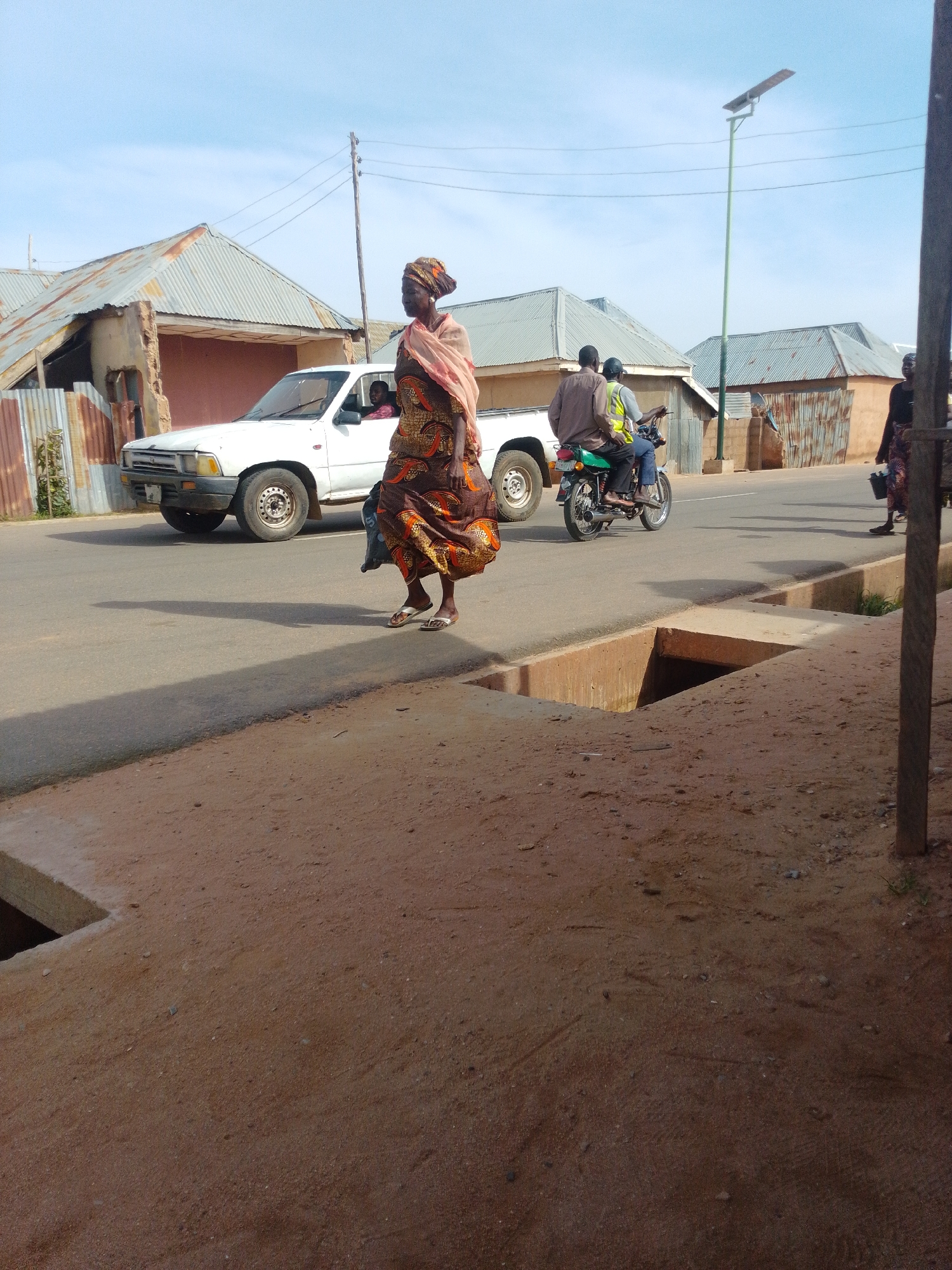 An exterior medium shot captures a street scene in Unknown, Unknown. A woman wearing a colorful patterned dress and a light pink headscarf walks across the road from left to right, her gaze directed forward. She is positioned in the foreground, her body angled towards the viewer. Behind her, a white pickup truck is parked, with a person visible in the driver's seat. Further in the background, a green motorcycle carries two individuals. The road surface is asphalt, with a concrete drainage ditch visible on the right side of the foreground. Several buildings with corrugated metal roofs are situated on either side of the street, some appearing weathered. A tall, slender pole with a solar-powered light fixture stands in the middle ground. The sky is a clear, light blue.