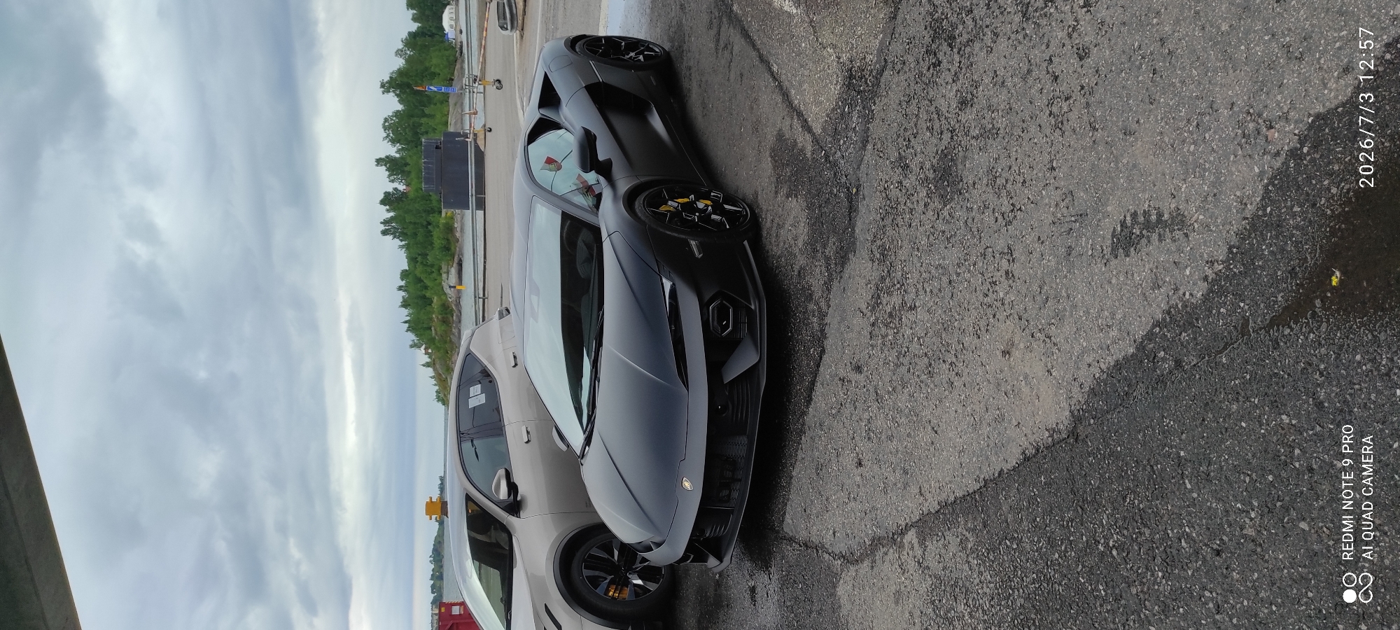 An outdoor, slightly overcast, daytime, eye-level shot shows two parked cars, a matte black Lamborghini and a silver SUV, on an asphalt surface next to a body of water. The Lamborghini, positioned in the foreground and angled to the right, is a low-profile sports car with yellow brake calipers visible through the spokes of its black wheels. The silver SUV is parked to the left of the Lamborghini and is partially obscured by it. In the background, across the water, is a dense line of green trees, and beyond that, a faint, cloudy sky. The water appears calm. The asphalt surface is wet and has some visible cracks and patches. A yellow bollard is visible on the left side of the background, near the water's edge. The image also contains watermarks in the bottom left and bottom right corners, indicating the device used for photography and the date and time.