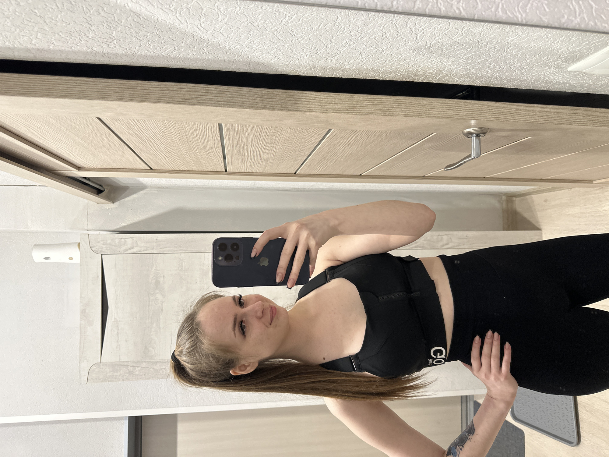 A woman is taking a selfie in an indoor setting. She is wearing a black sports bra and black leggings. The background includes a door, a cabinet, and a wall. The city and location are not identifiable.