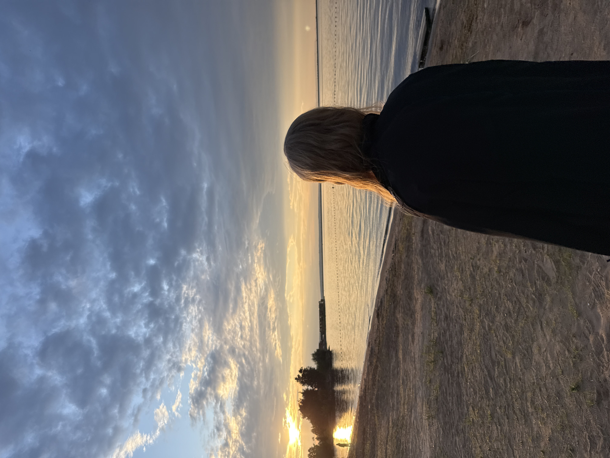 A person with blonde hair, facing away from the camera, stands on a sandy beach at sunset. The sun is low on the horizon, casting a warm glow on the water and the sky. The sky is filled with dramatic, textured clouds in shades of gray and orange. The water is calm, with gentle ripples reflecting the colors of the sunset. In the distance, there are trees and structures along the shoreline. The beach appears to be part of a park or recreational area, possibly on the Kama River in Chaikovsky, Russia. The overall atmosphere is peaceful and contemplative.
