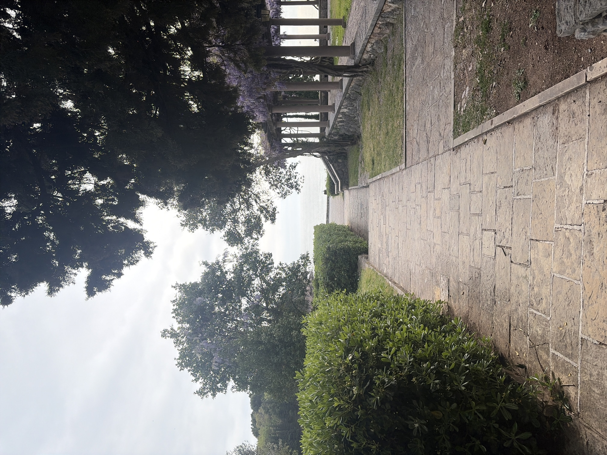 The image depicts a stone pathway leading towards a structure with columns, possibly a pergola, situated near the sea. The pathway is lined with greenery on either side, including manicured shrubs and trees. The sky is overcast. The image suggests a park or garden setting in an unknown city. There are no people visible in the image.