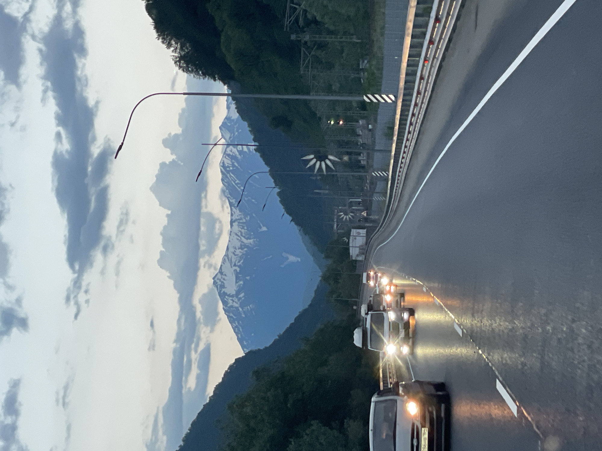 The image depicts a highway winding through a mountainous valley in Chvizhepse, Russia, during twilight or overcast conditions. The road surface is wet, reflecting the headlights of several vehicles traveling in the same direction. The vehicles include a silver sedan in the foreground on the left, and several vans and other cars further ahead, their headlights illuminated. The highway is bordered by a metal guardrail with reflective markers. Dense, dark green trees line the slopes of the mountains on either side of the valley, with the peaks of snow-capped mountains visible in the distance under a dramatic sky filled with dark, swirling clouds. Several streetlights with curved arms are positioned along the roadside. In the mid-ground, there are signs and what appear to be decorative wind sculptures. The scene suggests movement and travel along a scenic route.