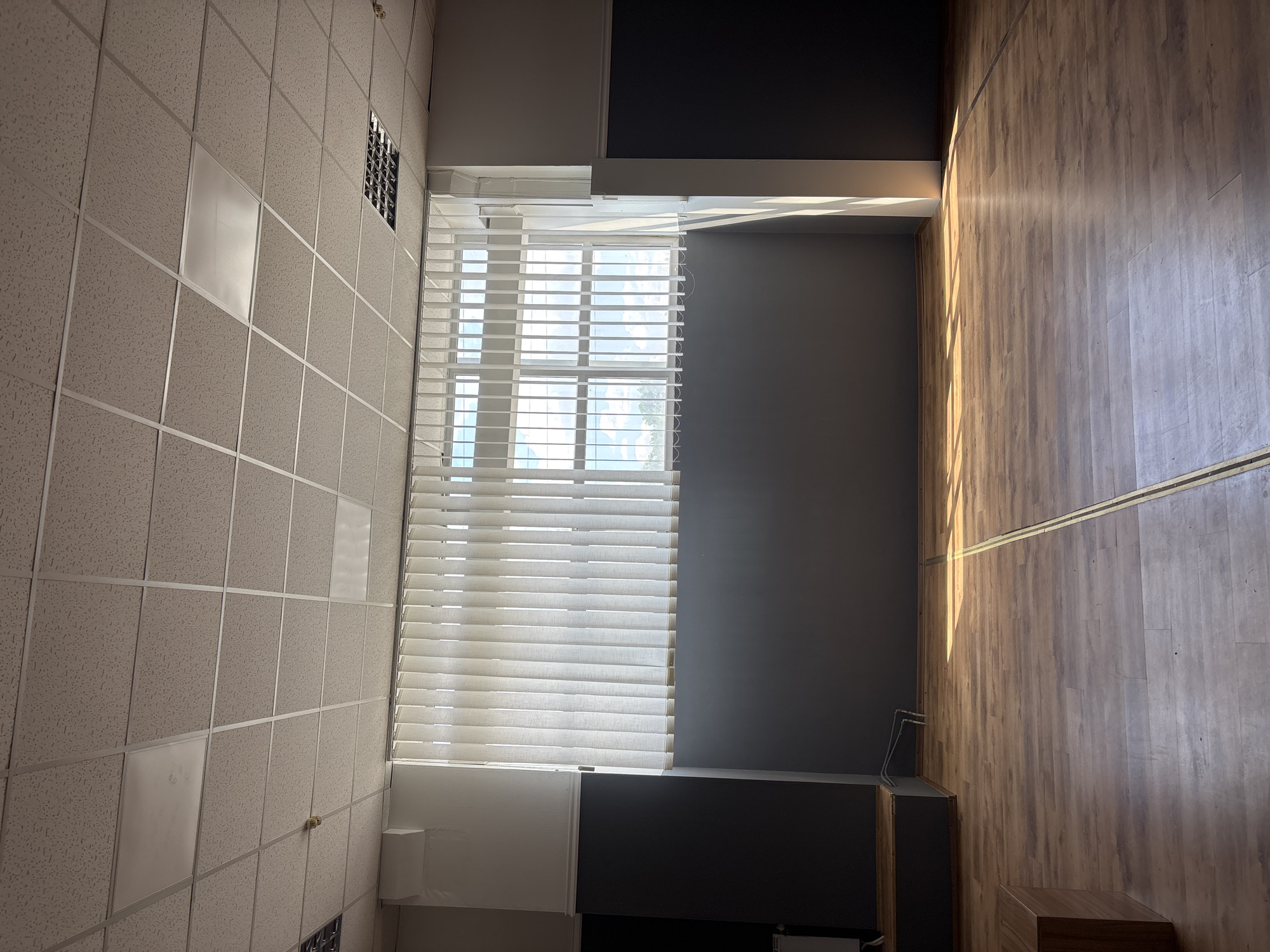 This is an interior photograph of an empty room, likely a studio or multipurpose space, located in Zaporizhzhia, Ukraine. The room has a light-colored, suspended acoustic tile ceiling with integrated fluorescent lighting fixtures and what appears to be a smoke detector. A large window spans a significant portion of the far wall, covered by vertical white blinds, through which bright daylight with scattered clouds is visible. The walls are painted in a two-tone scheme: a dark grey on the lower half and white on the upper half, separated by a molding. The floor is made of light brown laminate planks arranged horizontally. Natural sunlight streams in from the window, casting distinct linear shadows across the floor. In the foreground, to the left, a portion of a low, rectangular wooden bench or platform is visible. To the far left and right, dark panels or walls are present. Two thin, dark lines run vertically across the floor, suggesting a division or marking on the surface. No people or animals are present in the scene. The overall impression is one of an unoccupied, well-lit space.