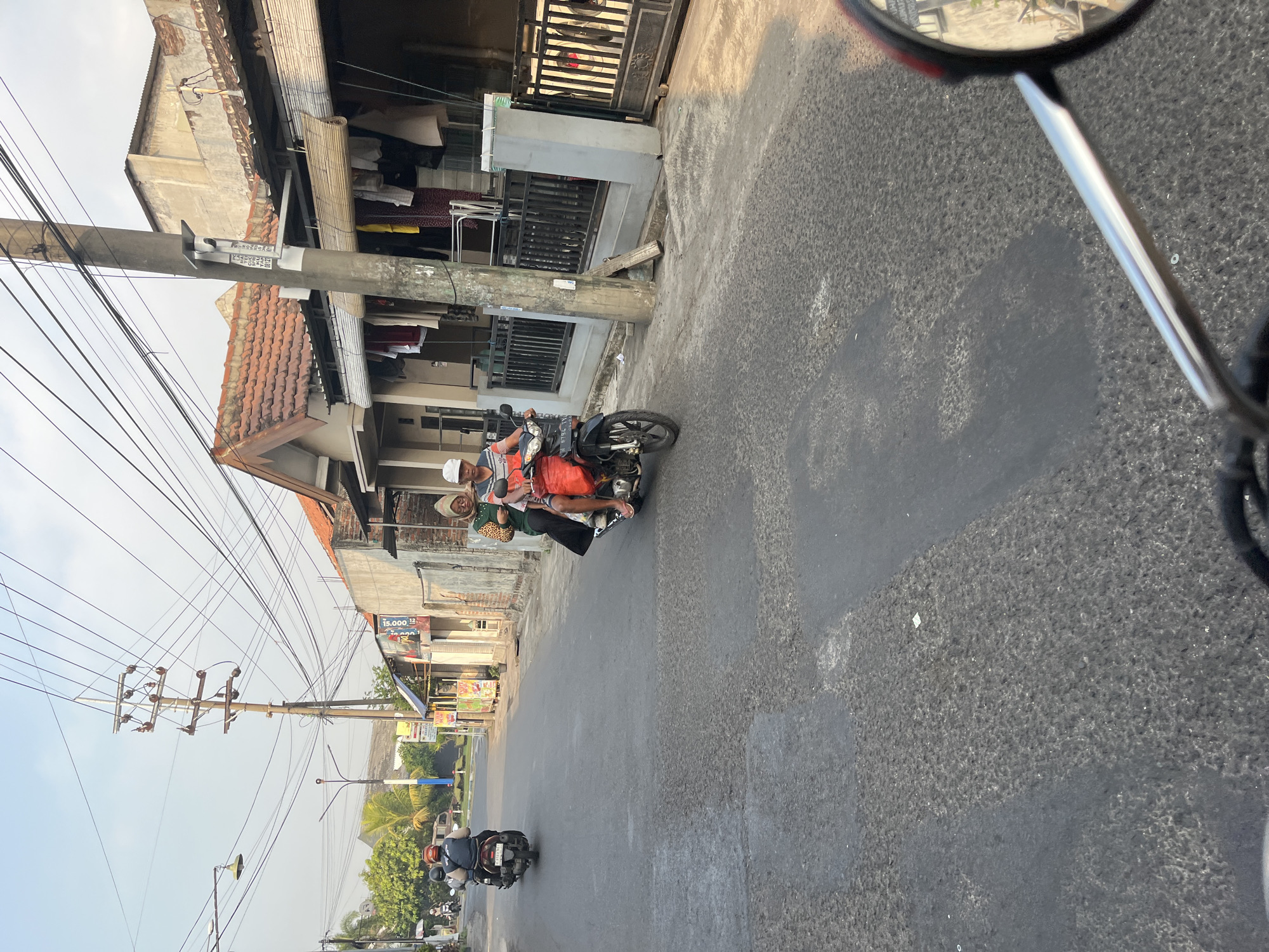 In Tenggulunan, Indonesia, two people are riding a motorcycle down a paved street. The person in front, wearing a white cap and a striped shirt, is driving. Behind them, a person wearing a hijab and a leopard-print shirt is the passenger. Both are looking forward. The street is lined with houses, some of which have tiled roofs and clothes hanging outside to dry. Utility poles with many wires are also visible. In the foreground, a motorcycle mirror reflects the scene.