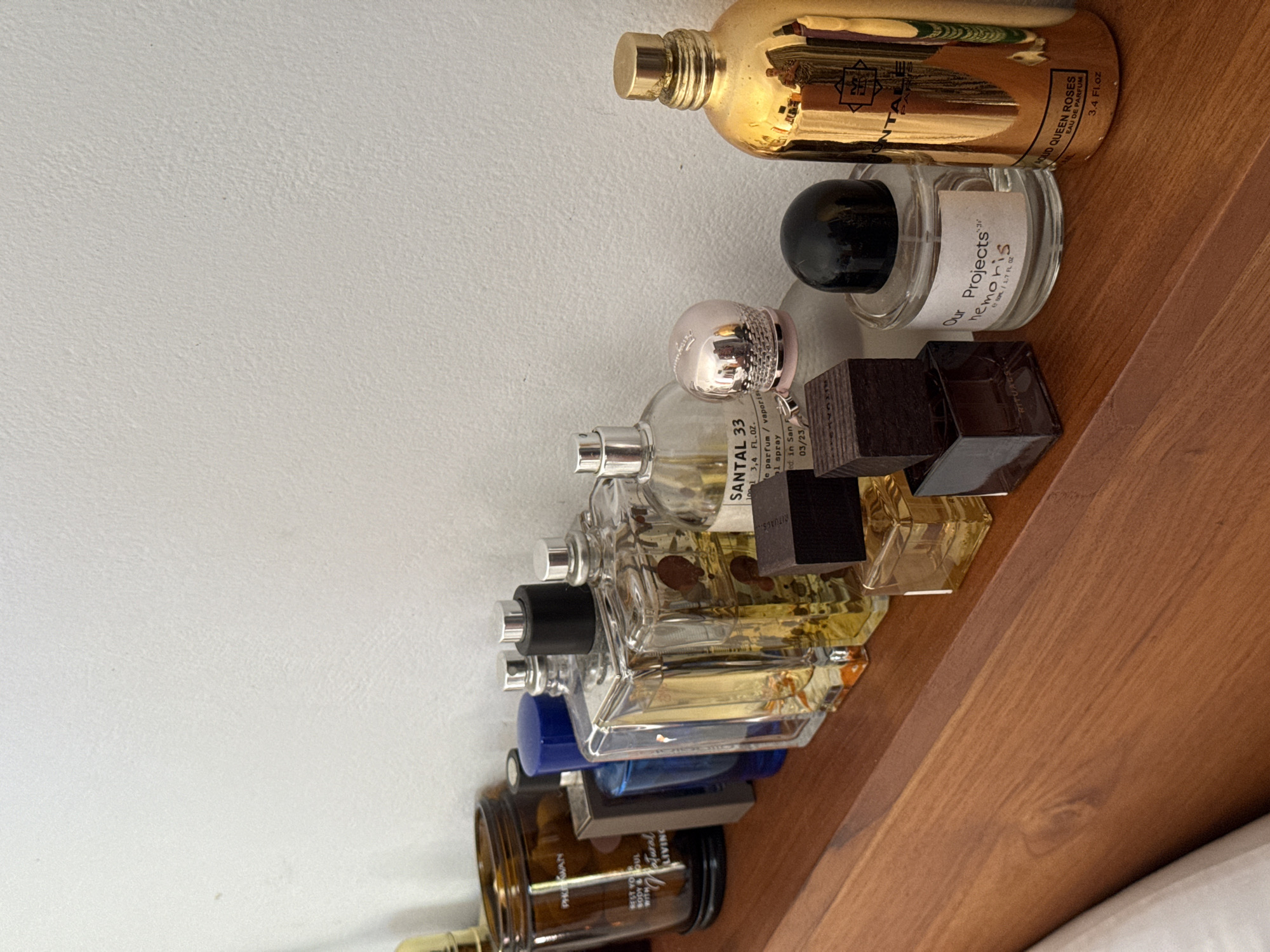A shelf holds a collection of perfume bottles. The top bottle is gold and labelled "AOUD QUEEN ROSES EAU DE PARFUM 3.4 Fl.oz". The next bottle is clear with a black cap and is labelled "Our Projects hemoris 50ML/1.7 FL OZ". The third bottle is clear with a pink cap and is labelled "SANTAL 33 1001 3,4 FL.OZ. e parfum / vaporisal spray ed. in San 03/23/". There are several more clear bottles below, along with a blue bottle and a brown bottle at the bottom of the shelf. The shelf is made of wood and is located against a white wall. There is no indication of the location in Pejeng, Indonesia.