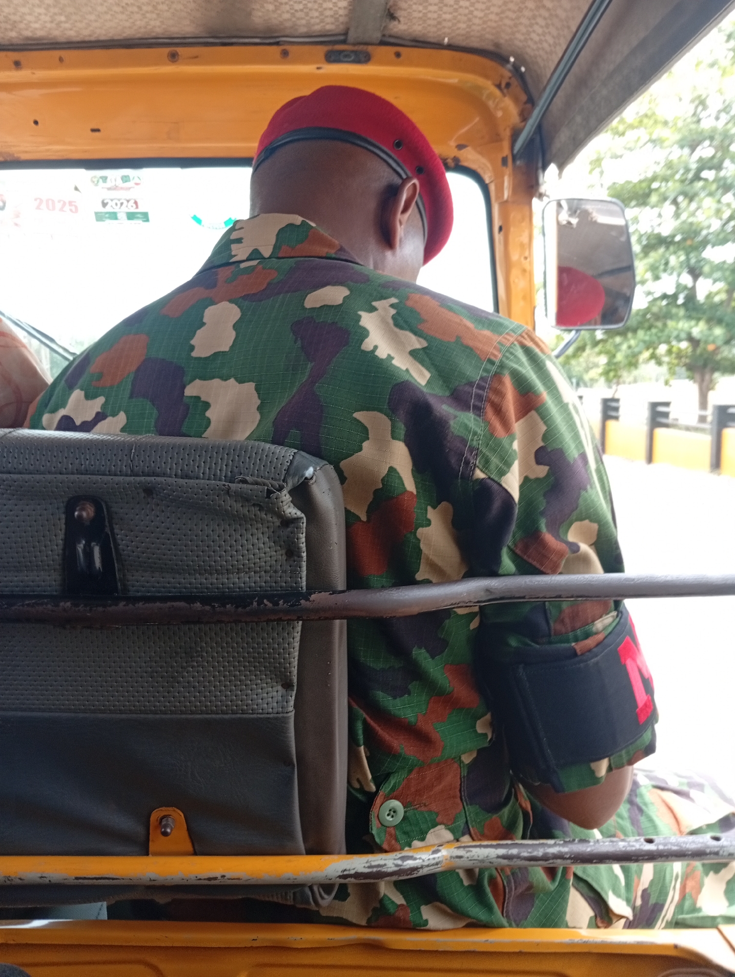 A medium shot, taken from behind, depicts a man in military attire riding in the back of a commercial tricycle, or "keke", in Adankolo, Nigeria. The man wears a red beret and a camouflage-patterned uniform. A black armband with a red "1" is visible on his left arm. He sits behind the driver's seat, positioned next to an old, torn, gray seat. The yellow interior of the tricycle is prominent. Through the tricycle's open window, a street scene is visible, including trees and a yellow and white barrier. Stickers displaying "2025" and "2026" are visible on the tricycle's front windshield.