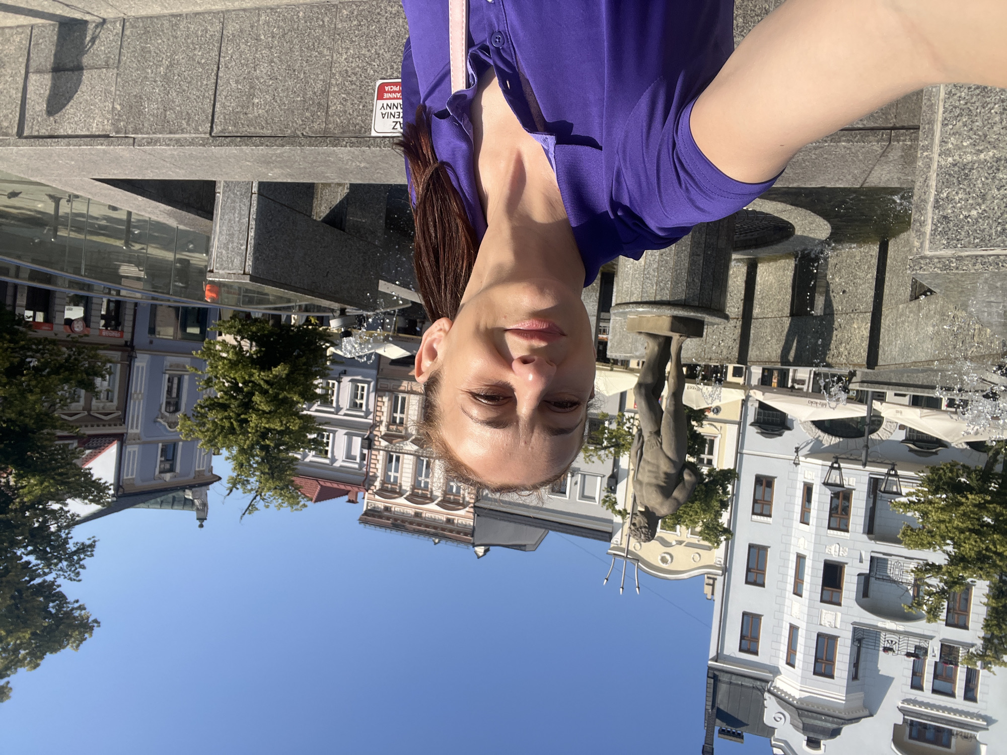 A woman with brown hair tied in a ponytail takes a selfie in front of a fountain and a statue of a man in a city square. The woman is wearing a purple polo shirt and a pink strap is visible over her shoulder. The fountain is made of granite and has water spraying. The statue is of a nude man holding a trident. In the background, there are several multi-story buildings with windows and balconies, and trees. The sky is clear and blue. The scene appears to be outdoors during the day.
