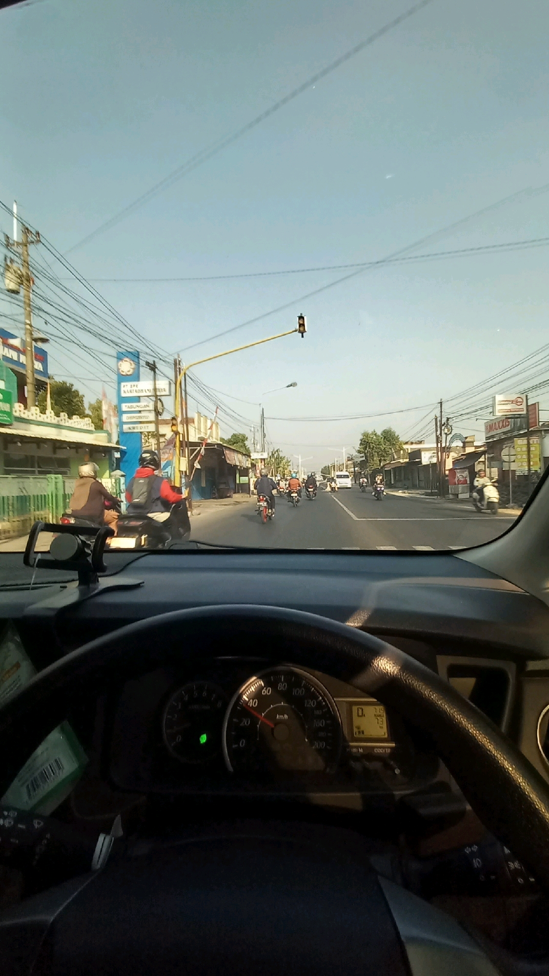 The image is taken from the perspective of a driver inside a vehicle, looking forward through the windshield. The scene depicts a street in Gentan, Indonesia, likely during the daytime, indicated by the clear blue sky and the bright sunlight casting shadows.
In the foreground, a portion of the vehicle's dashboard and steering wheel is visible. The speedometer shows the vehicle is traveling at approximately 20 km/h. There is a green indicator light illuminated on the dashboard, possibly for the turn signal or a similar function. A black car phone mount is attached to the dashboard.
The road ahead is lined with buildings and shops on both sides. A variety of vehicles, predominantly motorcycles, are on the road, with several individuals riding them. Some pedestrians are also visible on the sidewalks. Overhead, a complex network of electrical wires crisscrosses the sky, supported by utility poles. A traffic light is visible in the distance, suspended over the intersection. The overall environment suggests a typical urban or semi-urban street scene in Indonesia, characterized by moderate traffic and commercial activity.