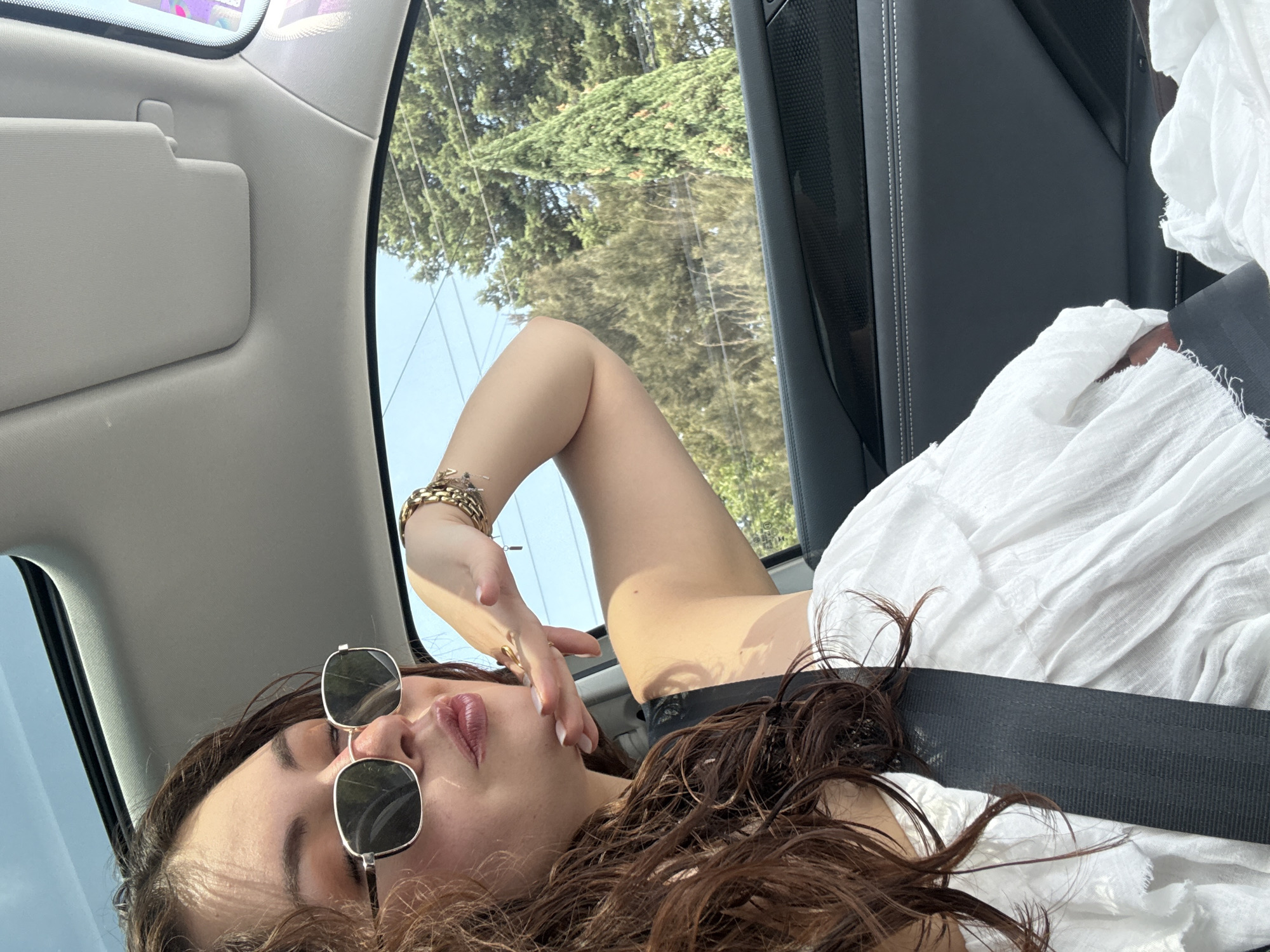 A female with brown hair and sunglasses is seated in the passenger seat of a vehicle, wearing a white garment and a seatbelt. Trees are visible through the car window. The location is unknown, and I am unable to confirm it is in Puebla, Mexico.
