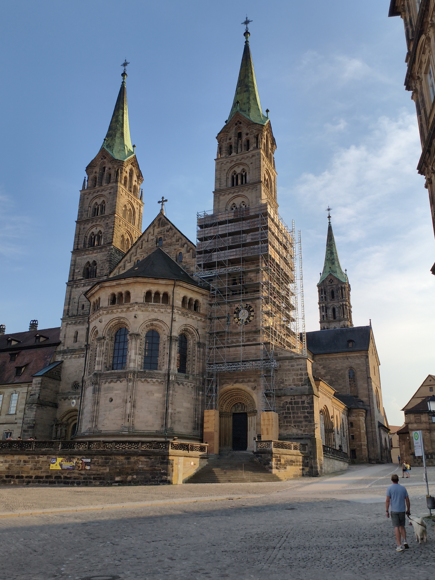 The image shows the Bamberg Cathedral, also known as the Imperial Cathedral of Saint Peter and Saint George, in Bamberg, Germany. The cathedral is a large Romanesque basilica with four towers, two of which are prominently visible in the foreground. The left tower is partially obscured by scaffolding, indicating ongoing renovations. A clock face is visible on this tower. The central portion of the cathedral has a large arched doorway. To the right, another tower with a green, pointed spire rises against a clear blue sky. The cathedral is constructed of stone, with intricate architectural details. In the foreground, a cobblestone plaza stretches out. A man and his dog are walking away from the camera on the plaza, towards the cathedral. Other people are visible in the distance, walking near the cathedral. The overall scene suggests a sunny day in a historic European city.