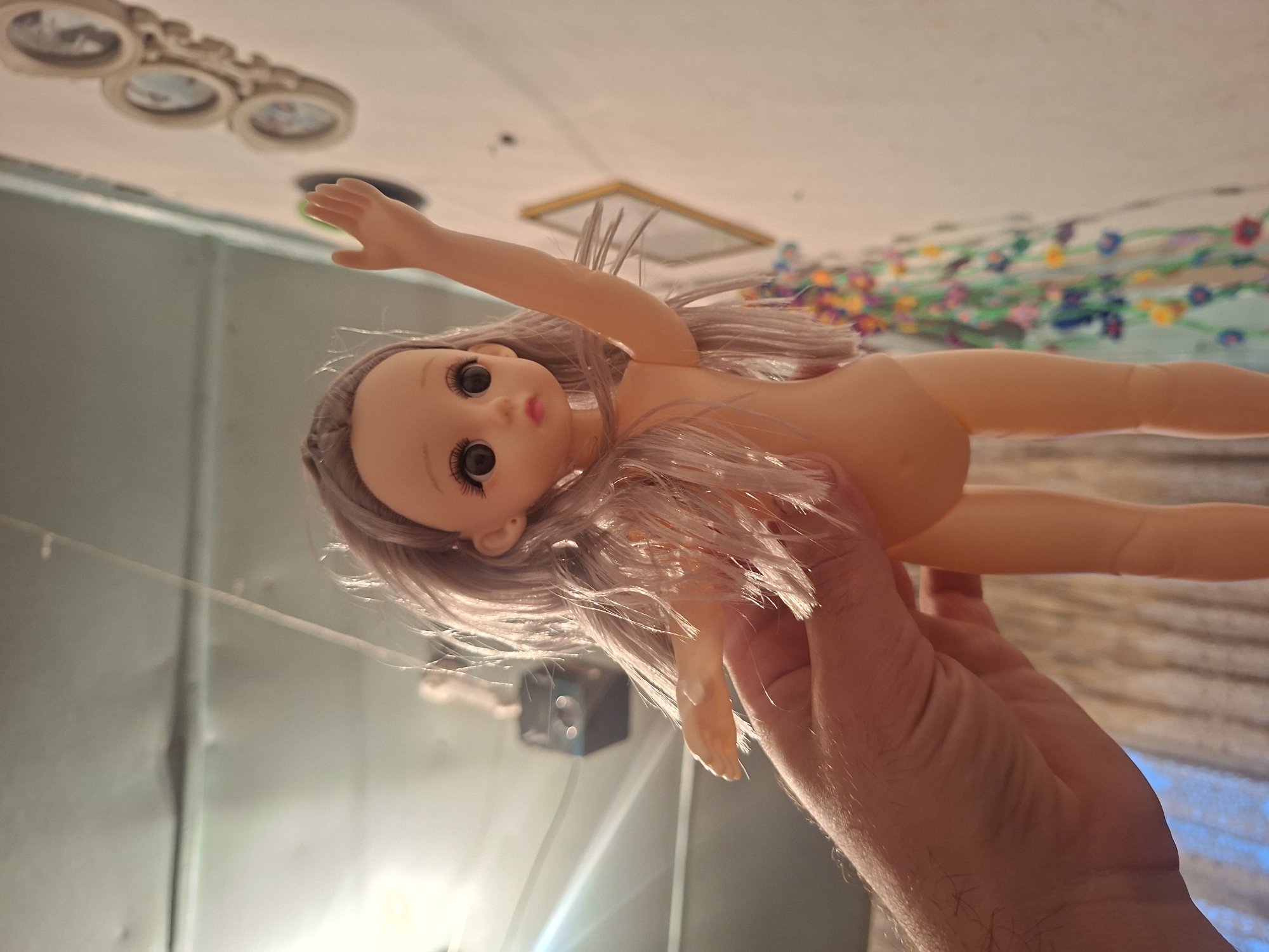 A medium, low-angle shot depicts a fair-skinned, naked doll being held by a human hand. The doll has gray hair and large, round eyes. The hand holding the doll is light-skinned and partially visible. In the background, the room's decor includes a wall, a ceiling light fixture, and a string of colorful decorations. The location is Unknown, Unknown.