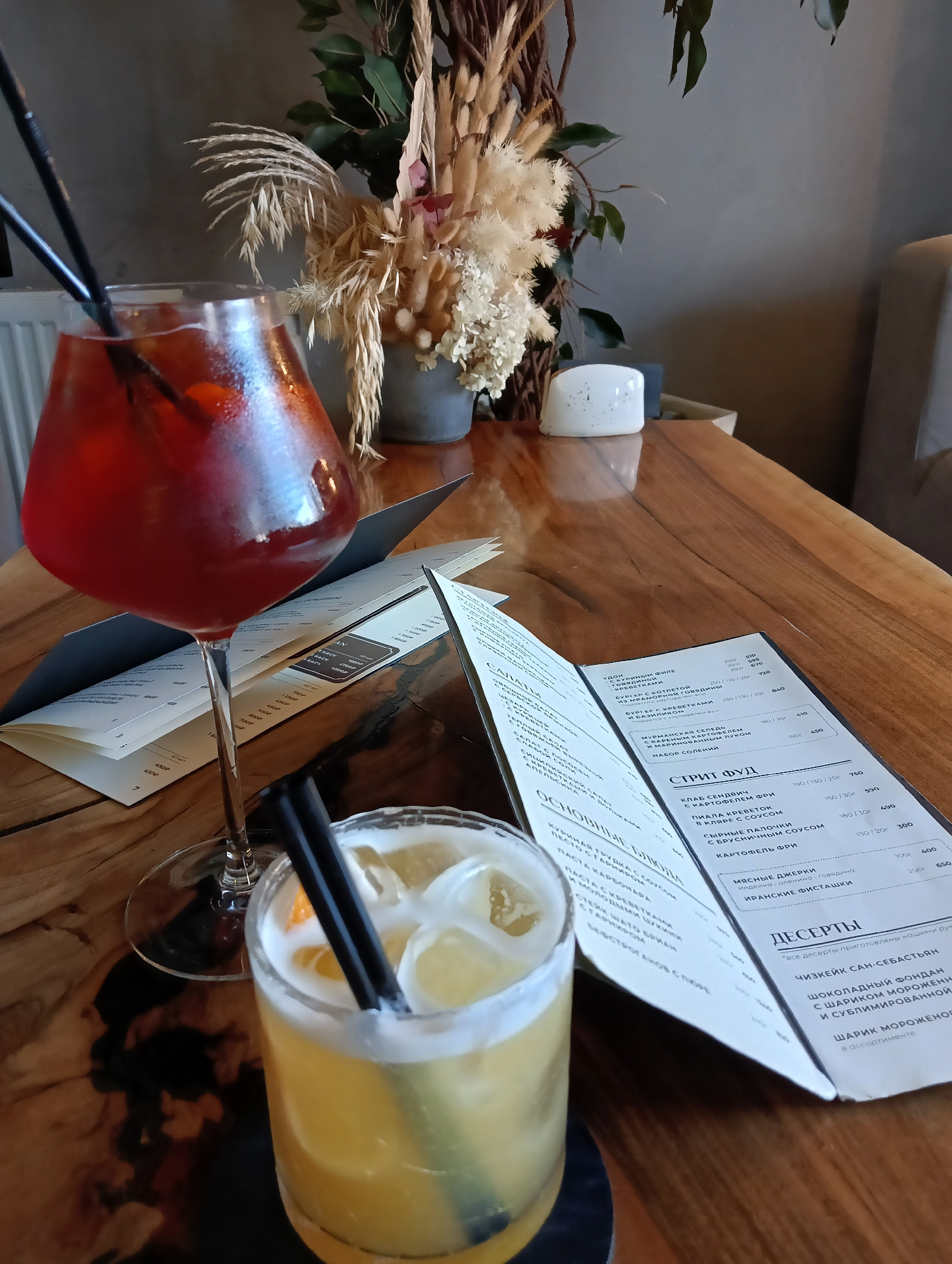 Two cocktails are placed on a wooden table in front of an open menu. The cocktail on the left is in a stemmed glass and is a deep red color with ice. The cocktail on the right is in a tumblr glass, is yellow, and has ice, a slice of orange, and two black straws. A bouquet of dried flowers and greenery is in the background. The menu is partially visible, displaying text in Russian, suggesting the location is Saint Petersburg, Russia. The setting appears to be a restaurant or cafe.